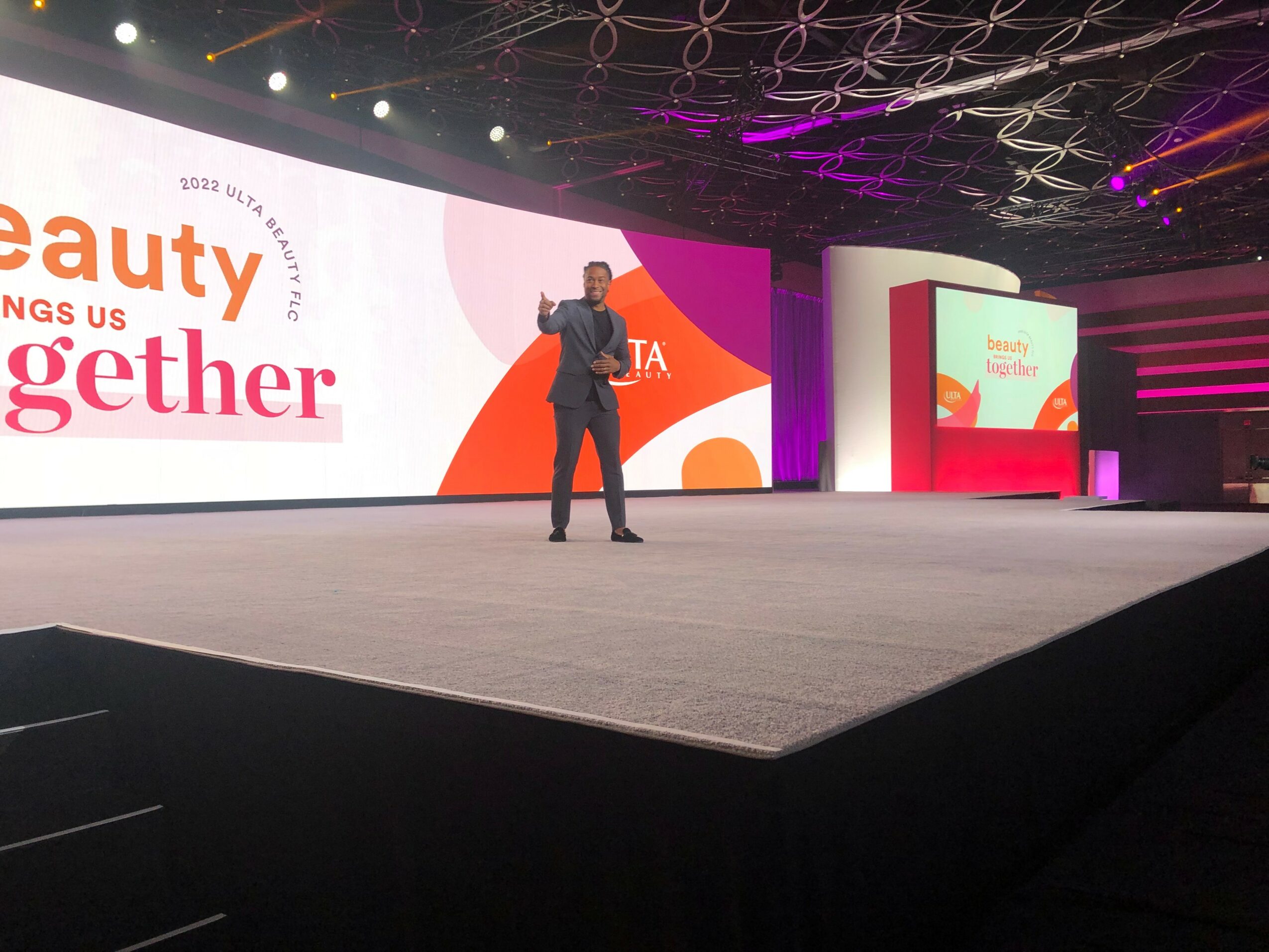 Person standing on stage with large screen behind, displaying text and graphics, in a conference or event setting.