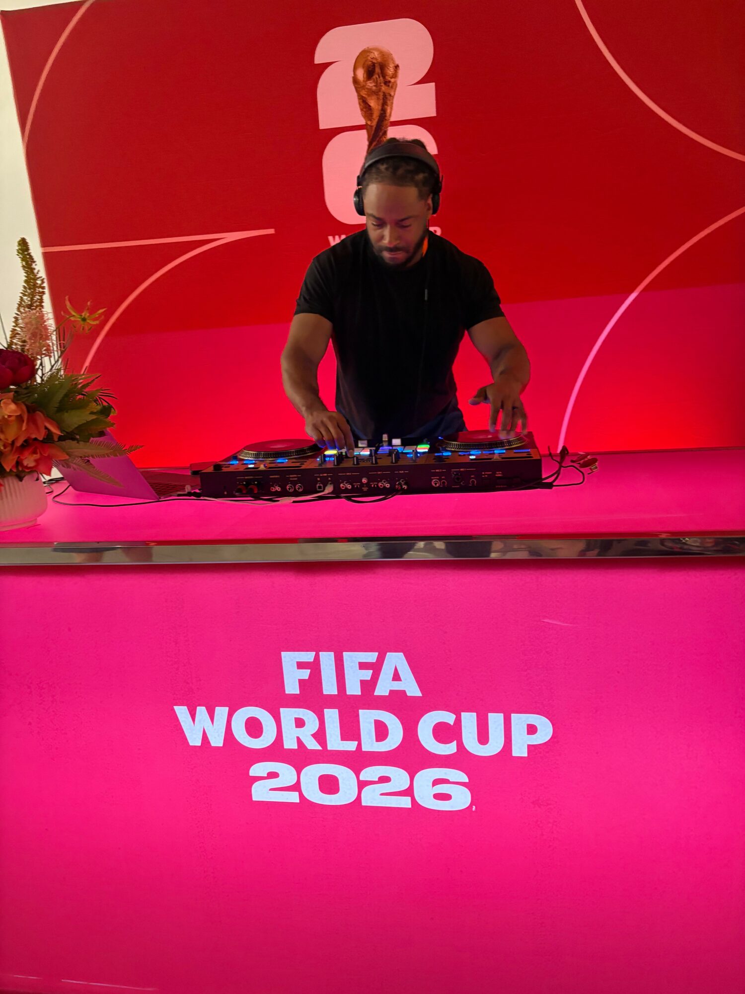 DJ performing at FIFA World Cup 2026 event with red background and FIFA logo, decorated with flowers.