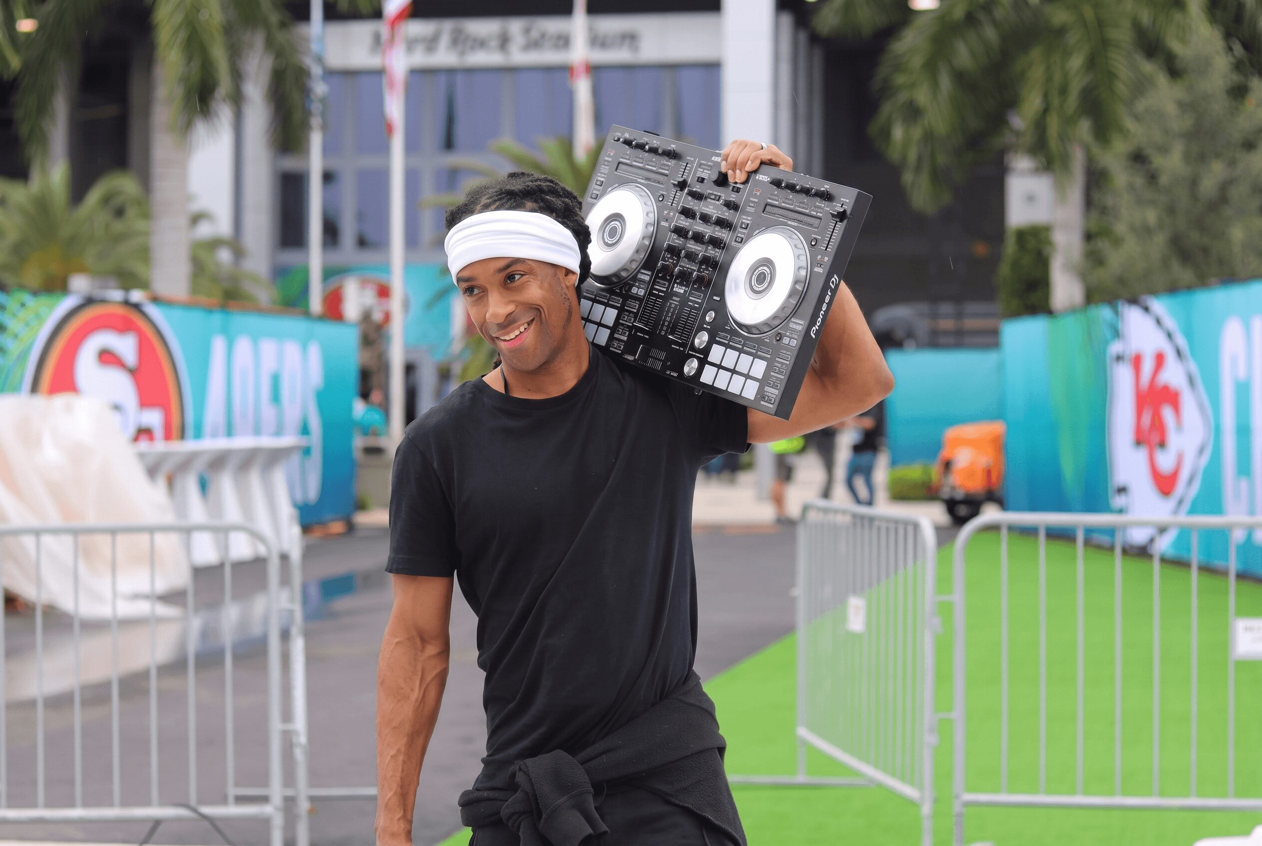 Man in black shirt and headband holding DJ equipment outdoors, with barriers and colorful banners in background.