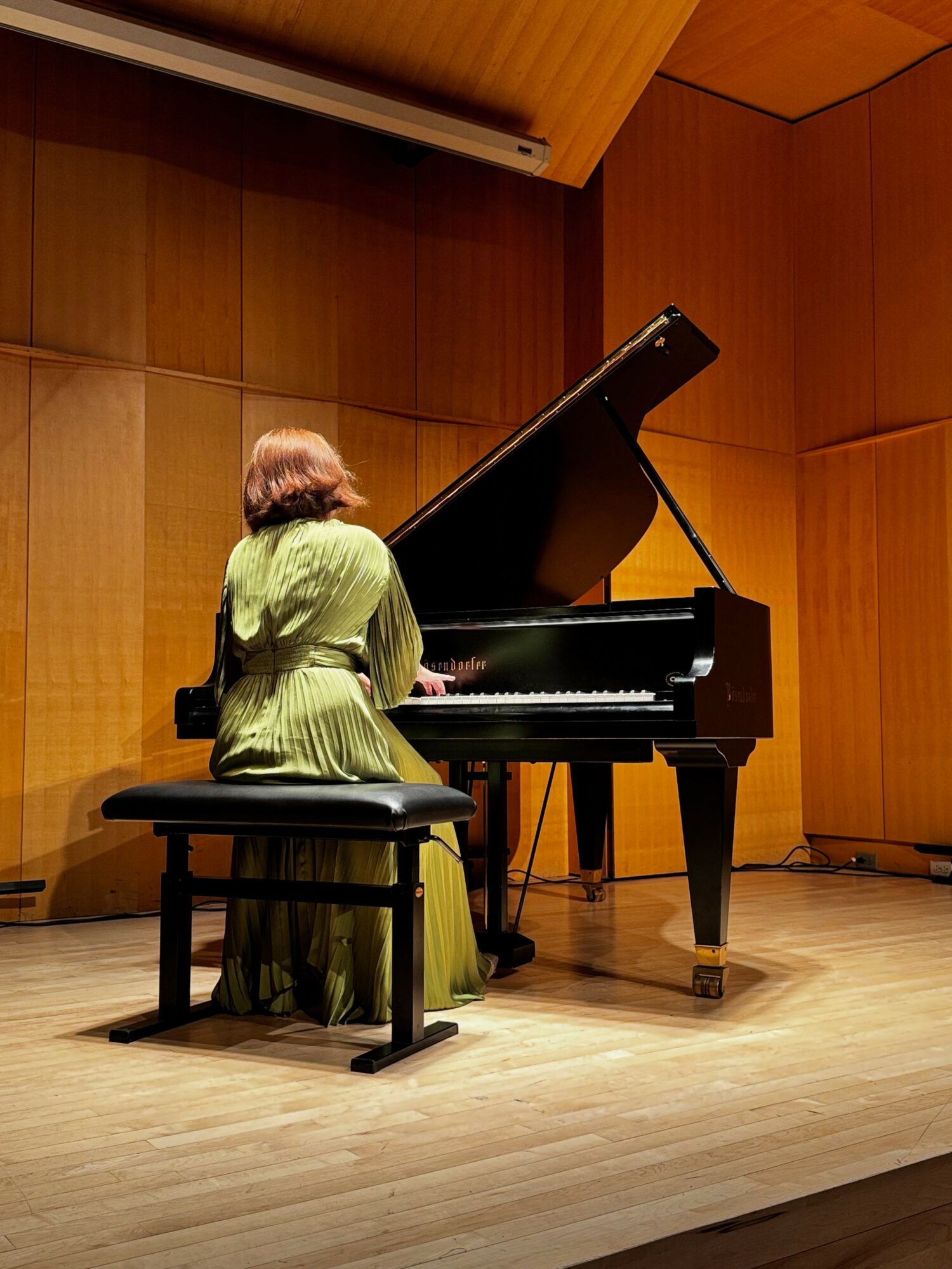 Person with short hair in a green dress playing a grand piano on stage with wooden walls.