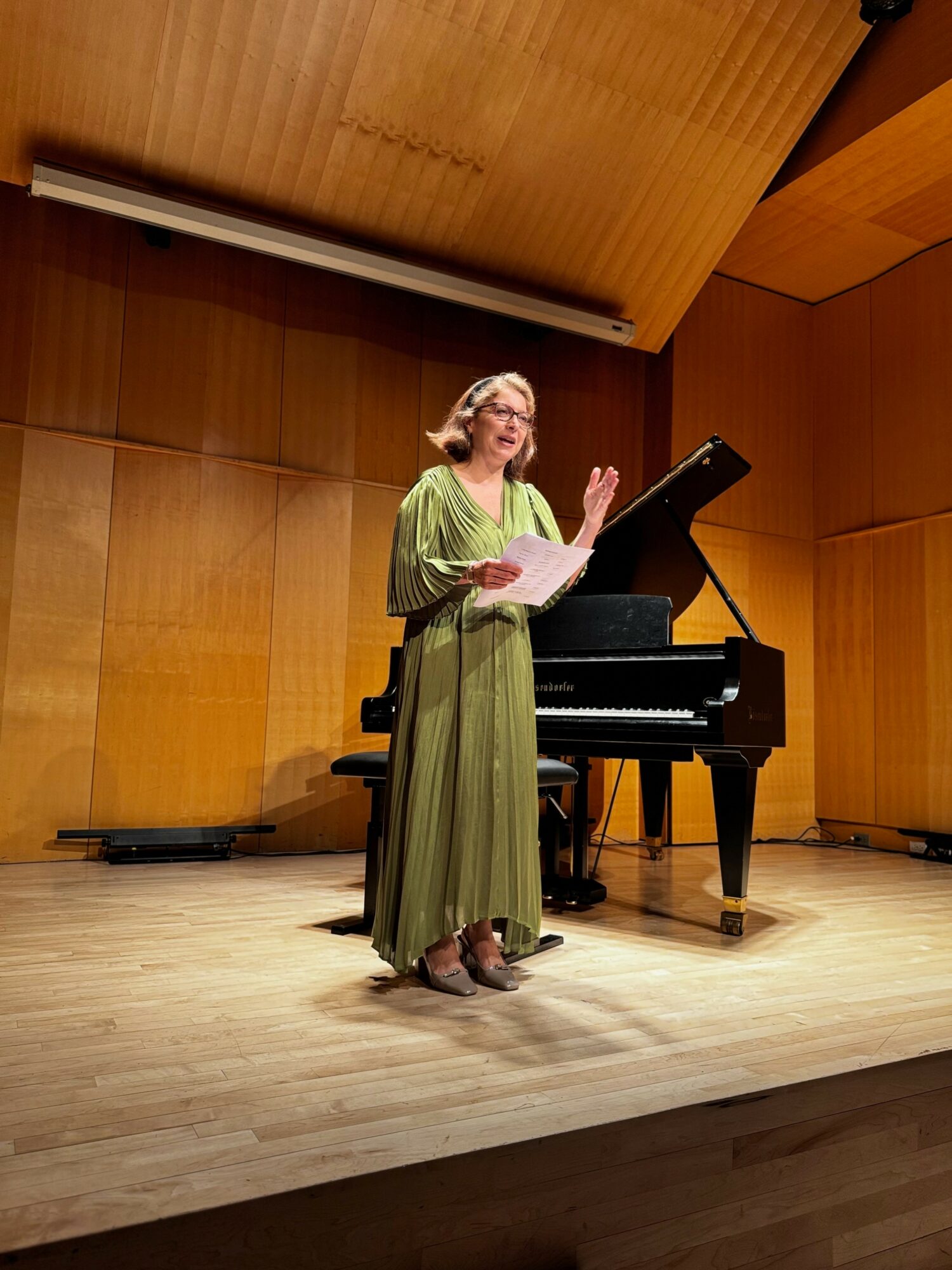 Woman in green dress singing on stage with a grand piano, wooden walls, and ceiling, stage lighting, and sheet music.
