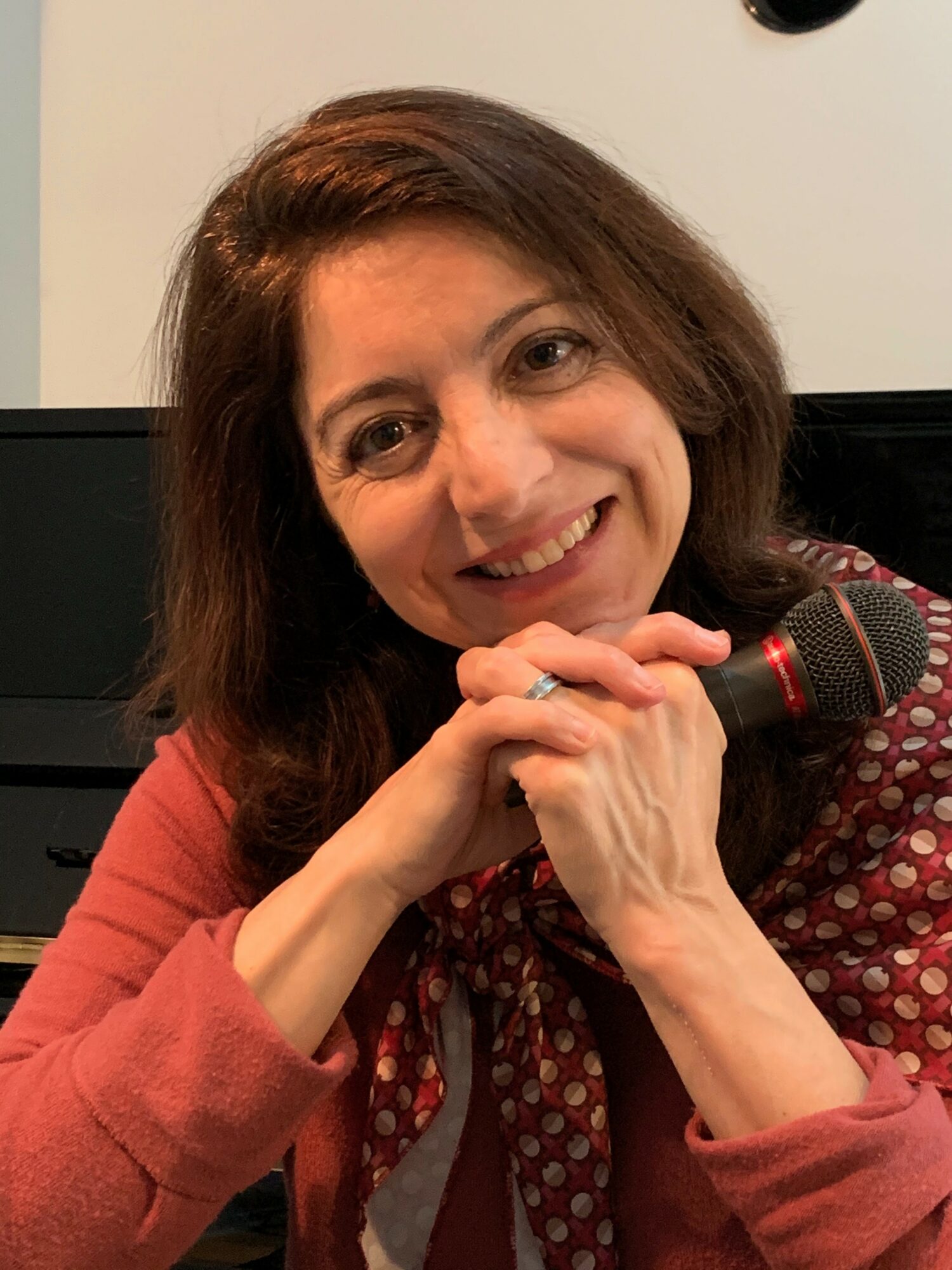 Woman with brown hair smiling, holding a microphone, wearing a patterned scarf and a reddish top.