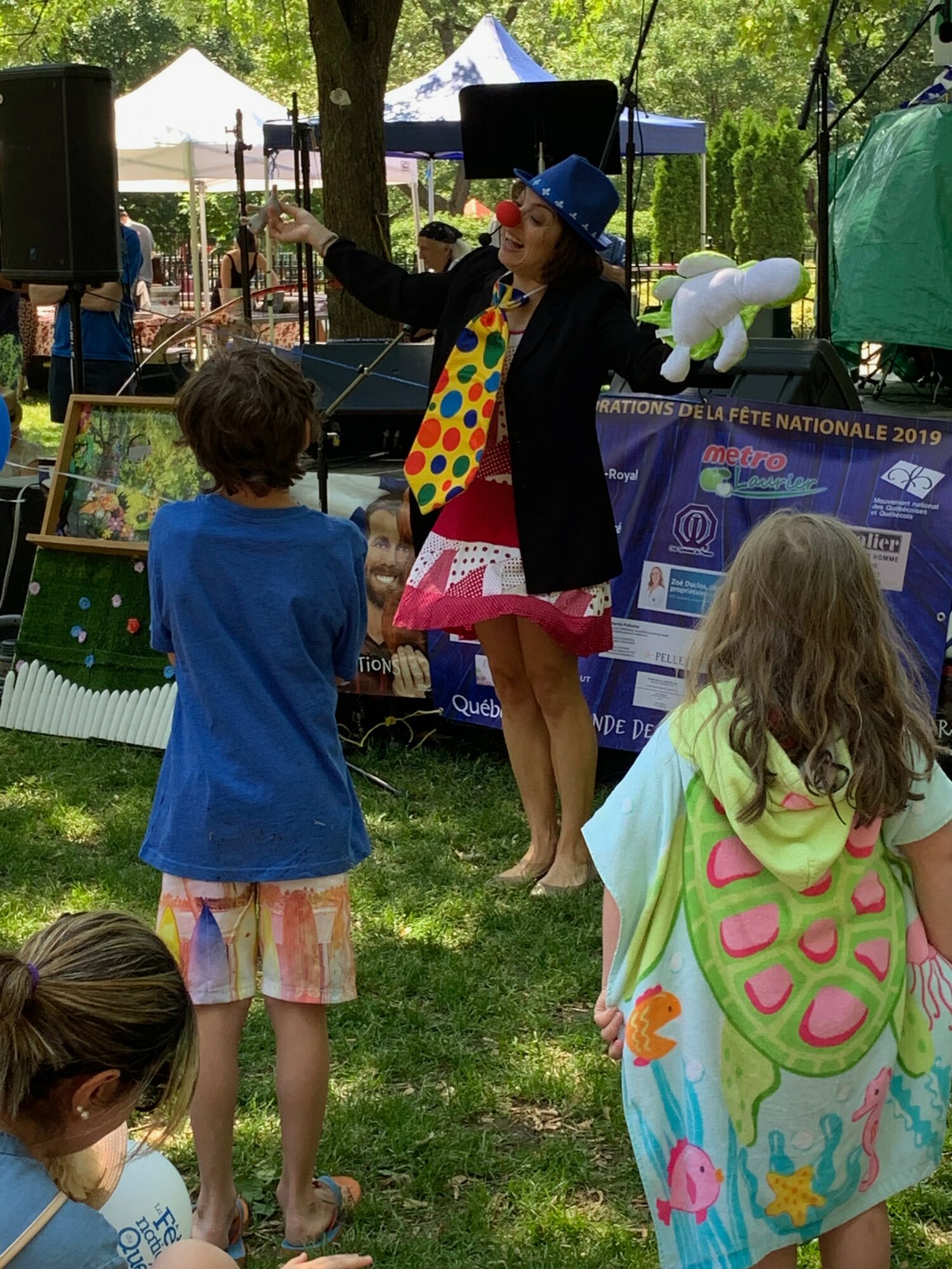 A woman in a blue hat and colorful dress performs a puppet show for children outdoors under trees, with tents in background.