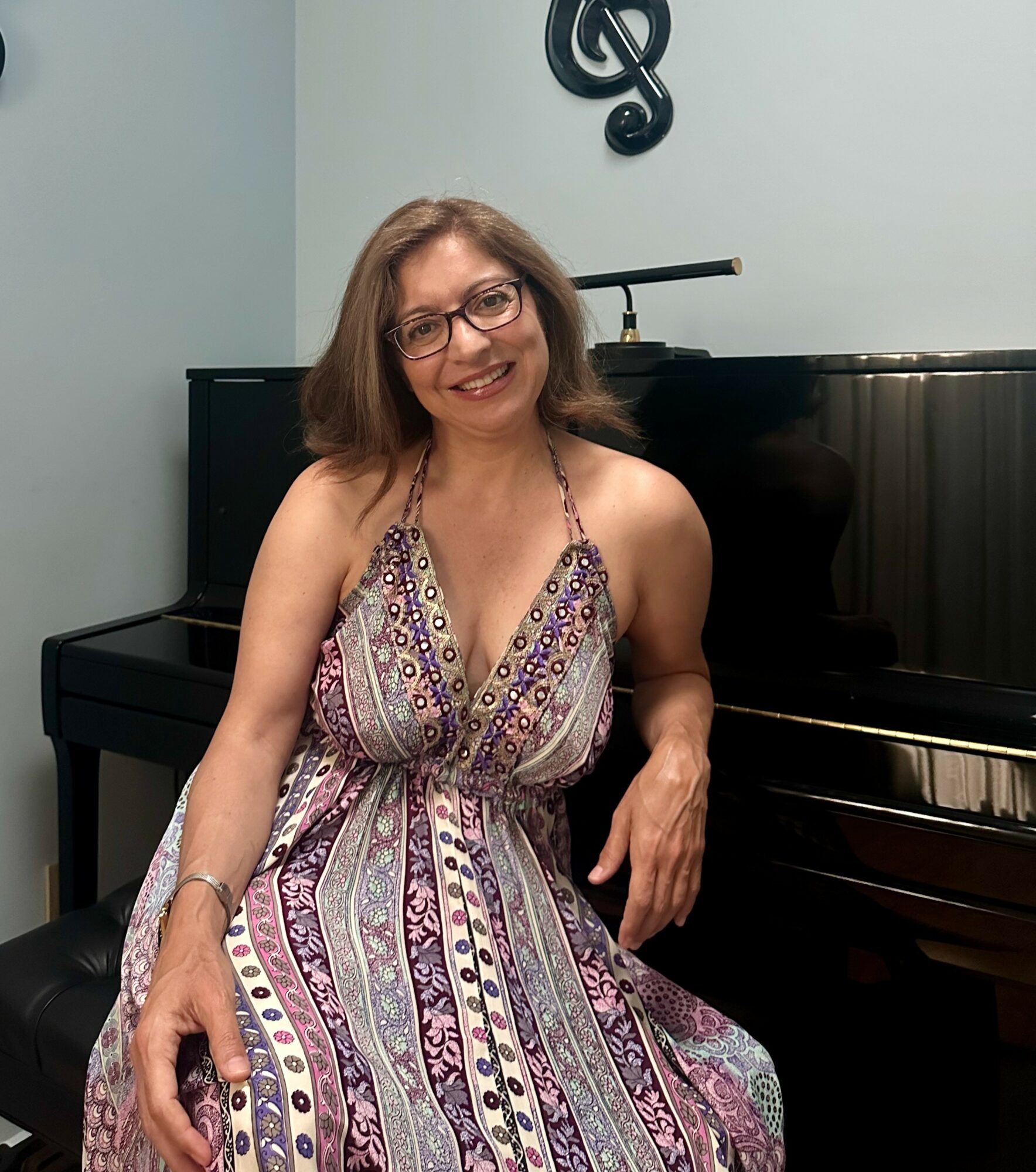 Woman with glasses in a patterned dress sitting beside a piano, smiling, in a room with a musical note decoration on the wall.