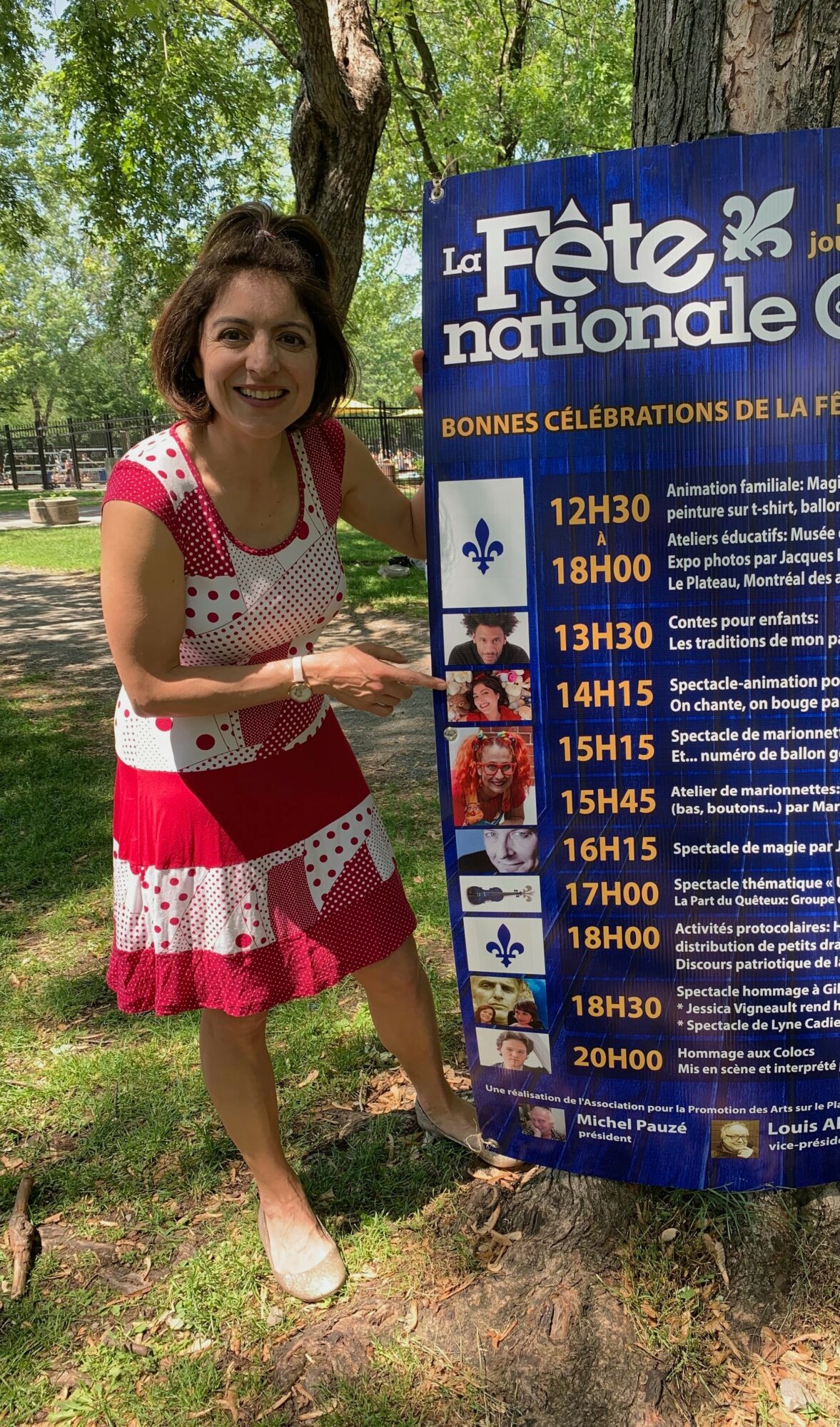 Woman standing outdoors next to a large blue event schedule sign, smiling, trees in background.