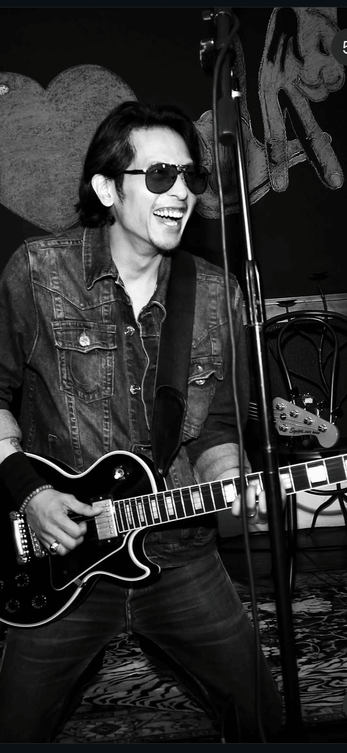 Person playing guitar, smiling, wearing sunglasses and a denim jacket, performing on stage with microphone and equipment.