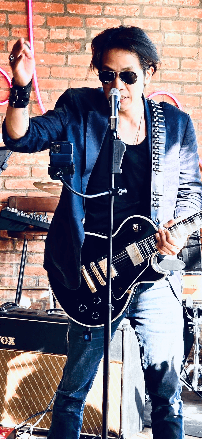 Musician with sunglasses, black guitar, and dark jacket performing on stage against a brick wall.