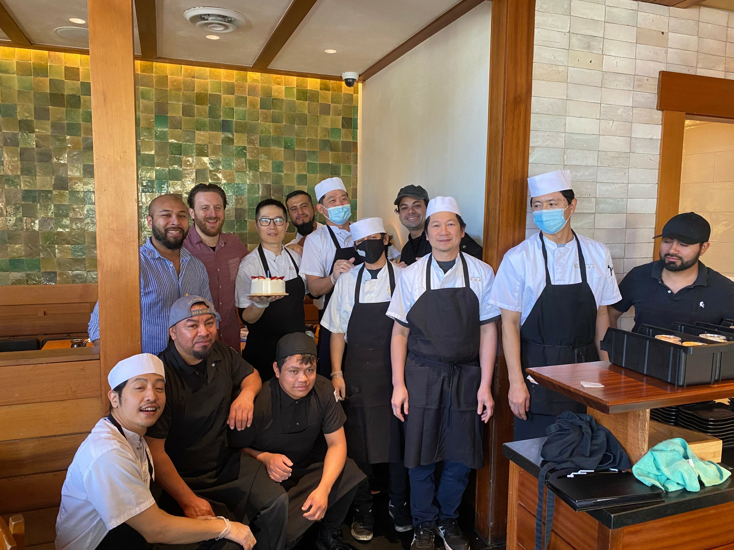 Group of people, some in chef uniforms and masks, gathered in a restaurant with colorful tiled wall, smiling and posing.