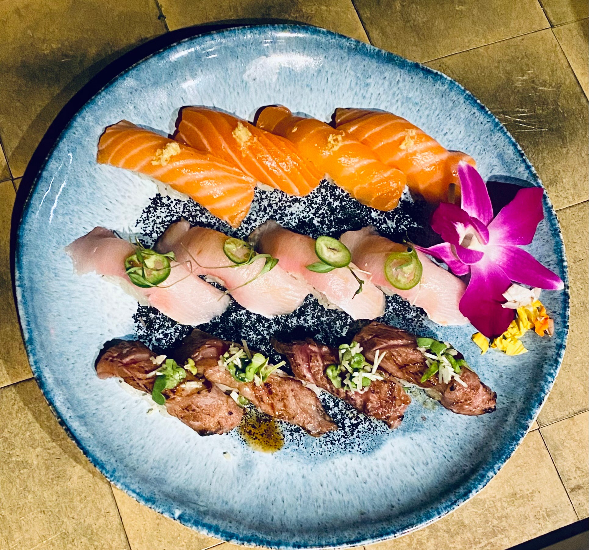 Assorted sushi with salmon, tuna, and beef, garnished with cucumber slices and a purple orchid flower on a blue plate.