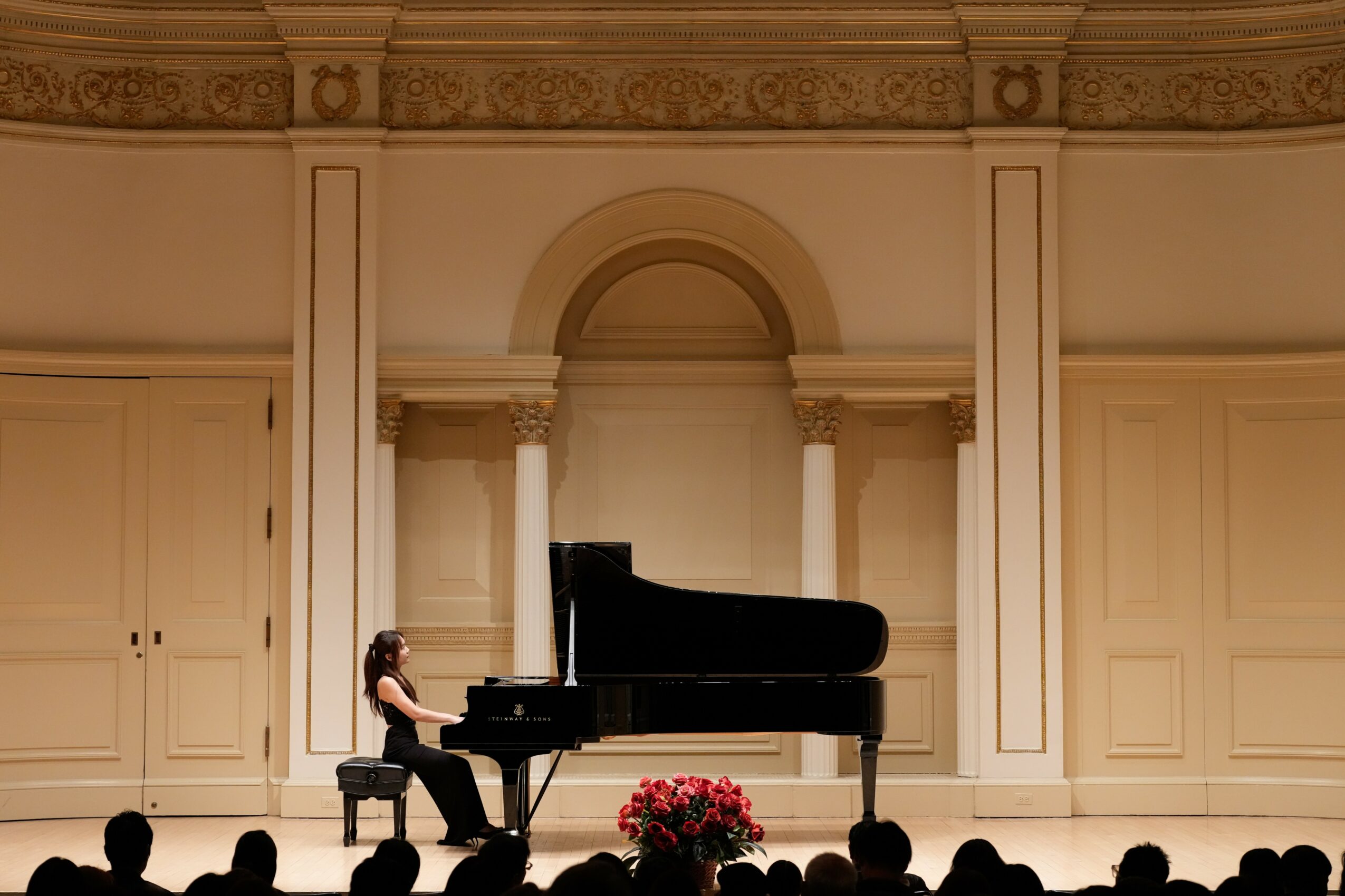 Person playing piano on stage with floral arrangement in front, ornate background, and stage lighting, no visible audience.