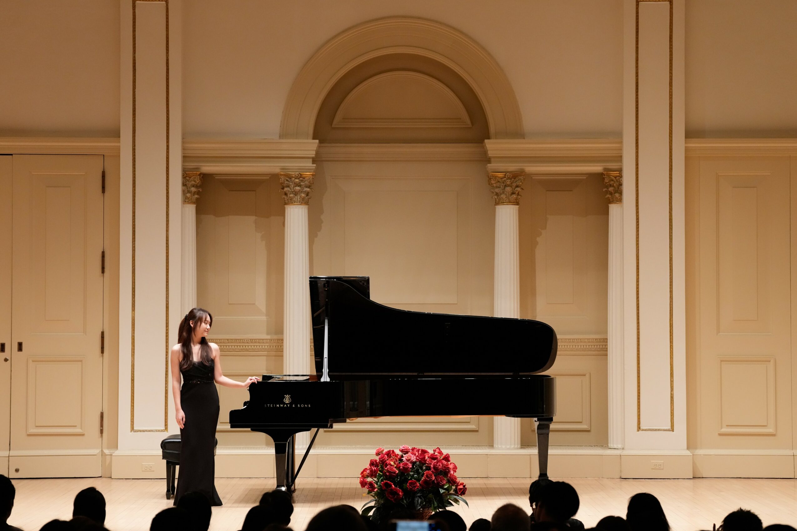 A woman stands on stage near a grand piano with a bouquet of flowers in front, in a classical concert hall.
