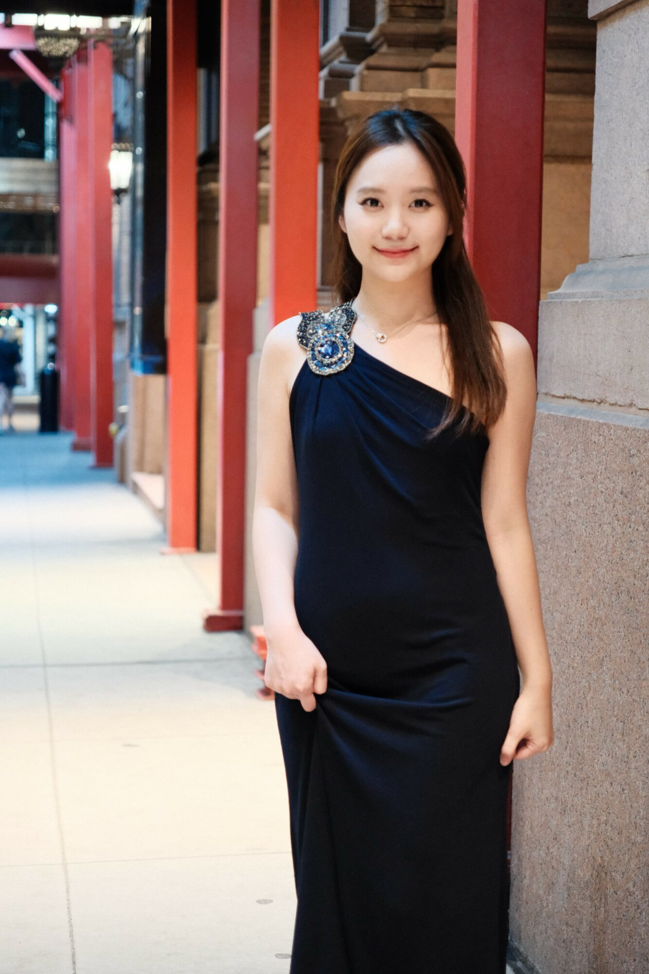 Young woman in a black dress standing outdoors near a building with red columns and stone wall.