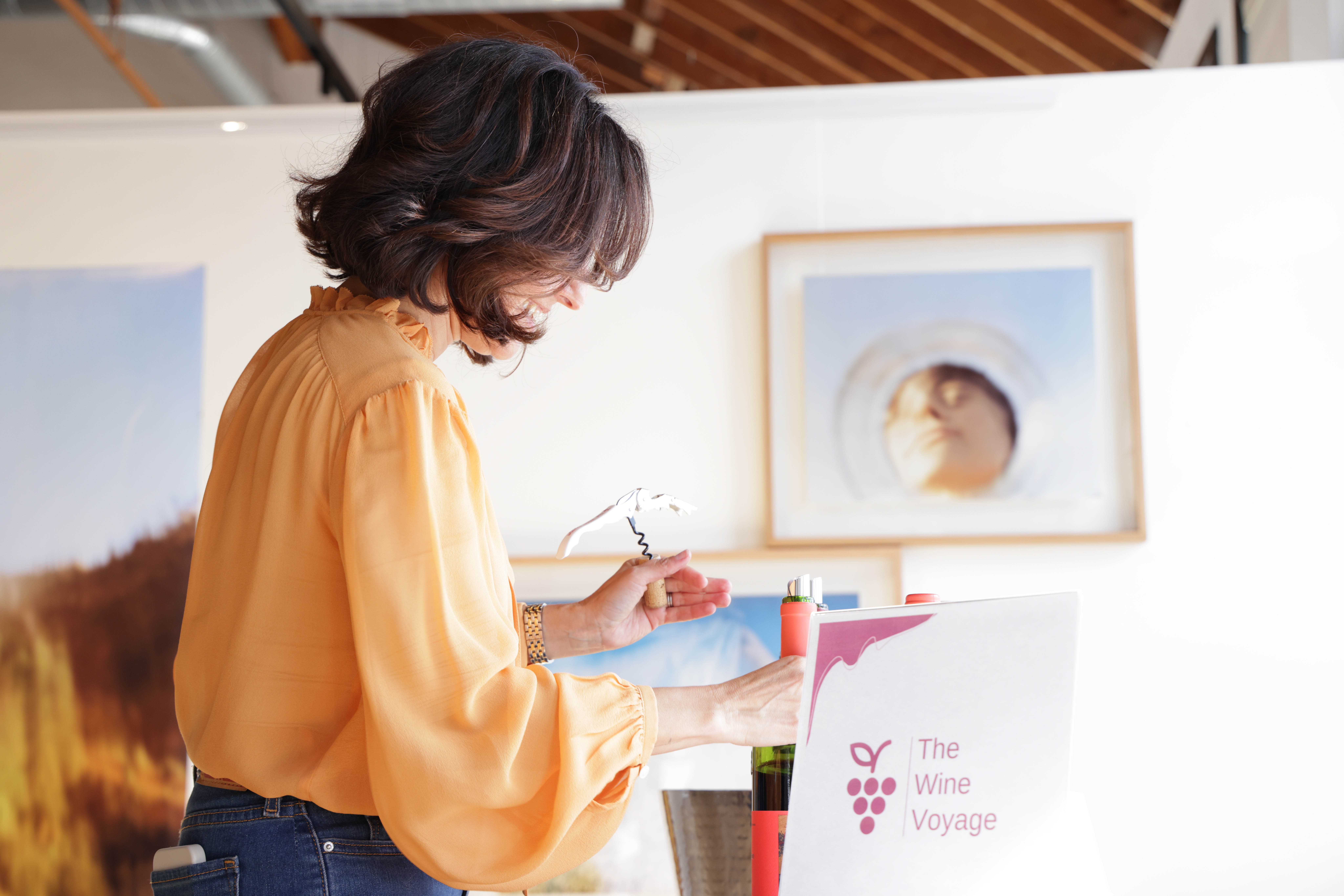 Woman with dark hair in an orange blouse looks at artwork in gallery, holding a glass of wine.