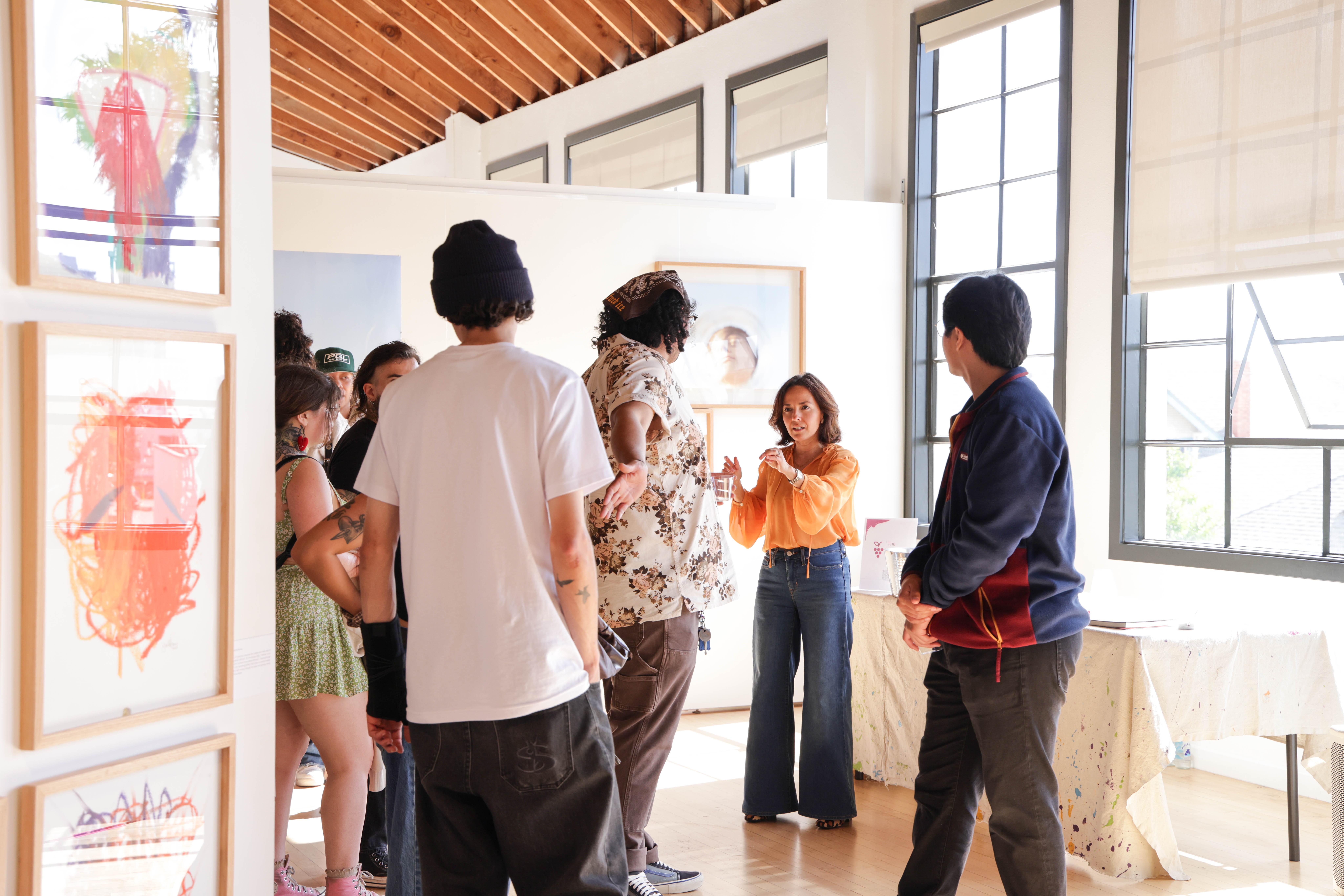 Group of people in an art gallery, some looking at artwork and others listening to a woman speaking, bright room with large windows.