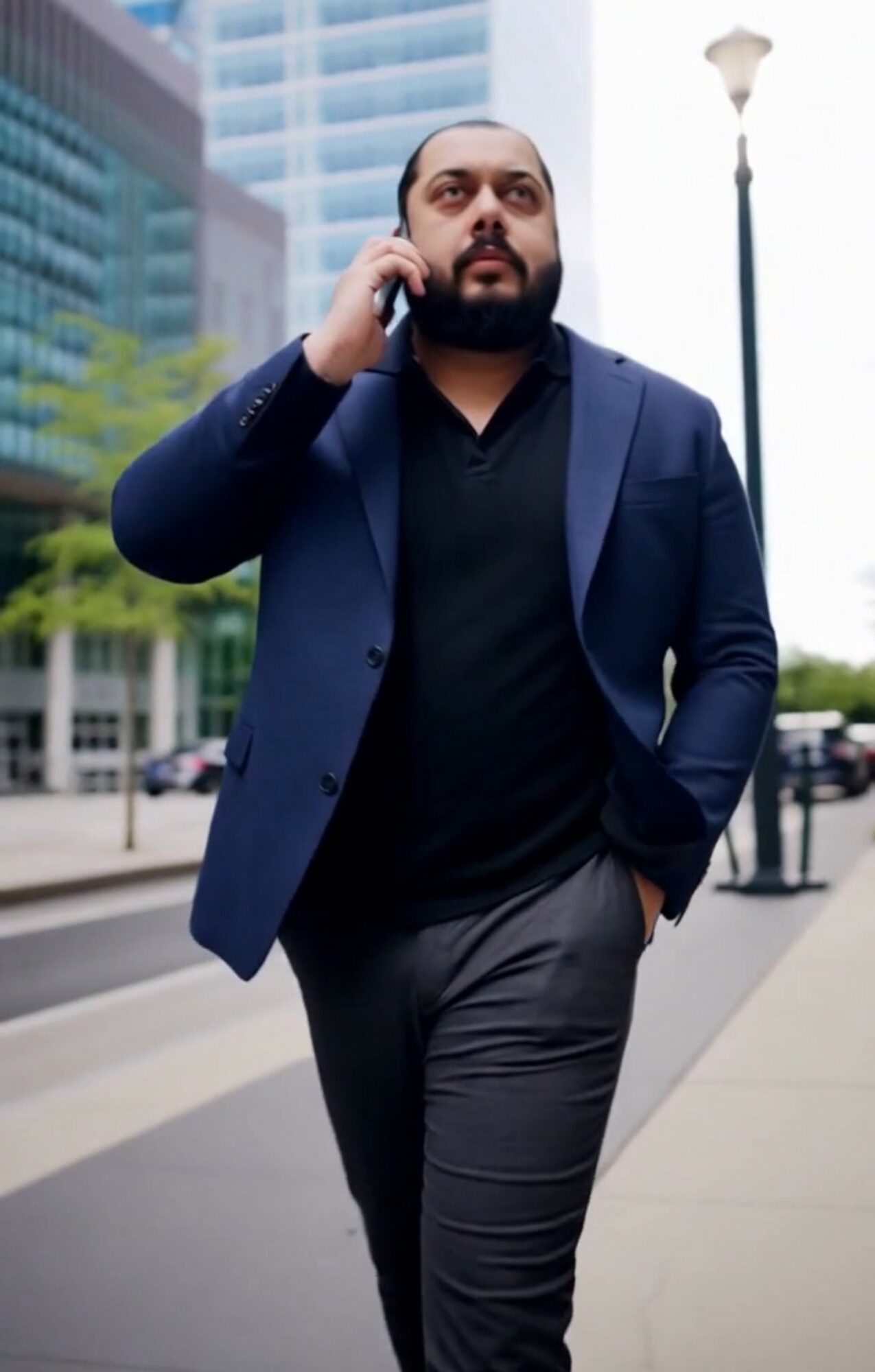 Man in business attire walking on city sidewalk, talking on phone, with modern buildings and streetlamp in background.