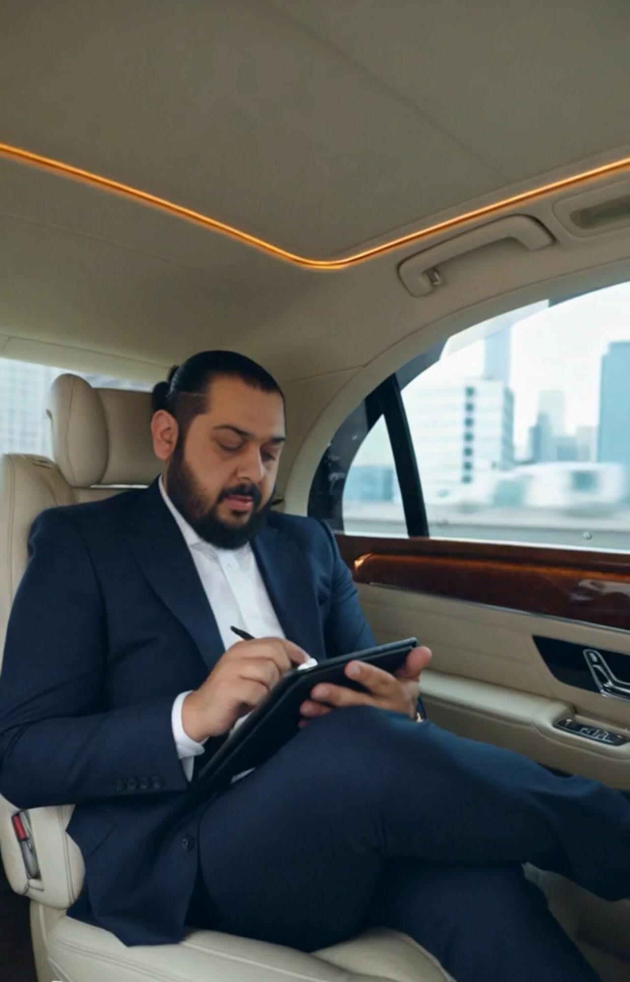 Man in suit sitting in car, using a tablet, city buildings visible outside window.