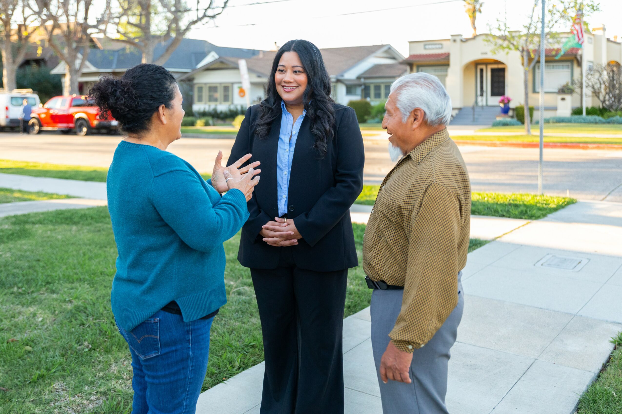 Three people, two women and one man, are talking outdoors on a sidewalk with houses in the background.