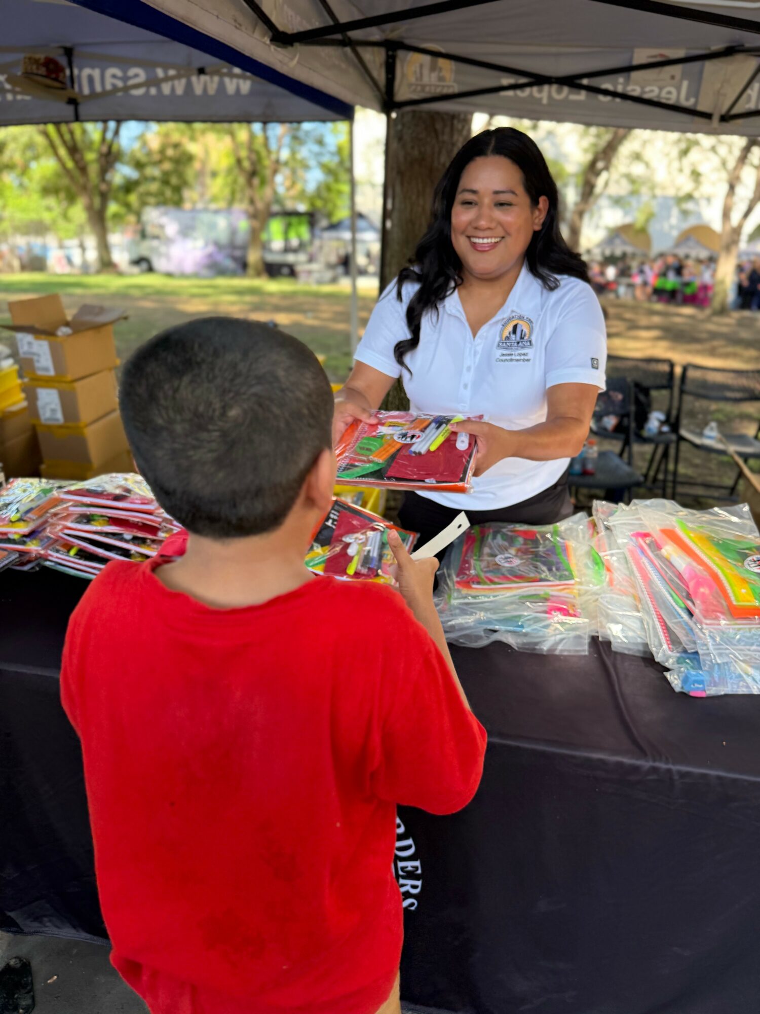 Woman smiling at a young boy at an outdoor booth with school supplies, trees in background.