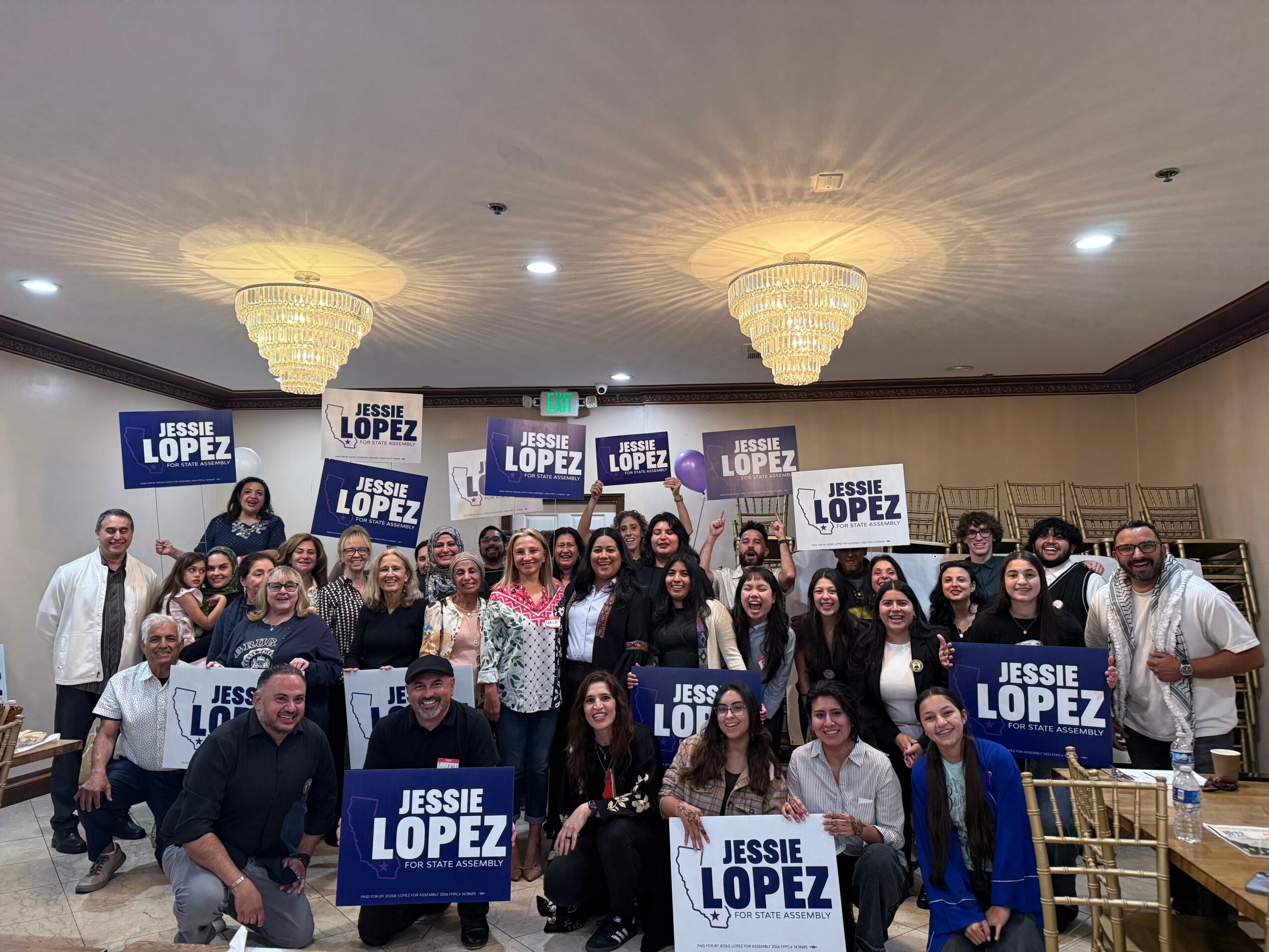 Group of people holding signs with candidate's name, posing indoors with chandeliers overhead.