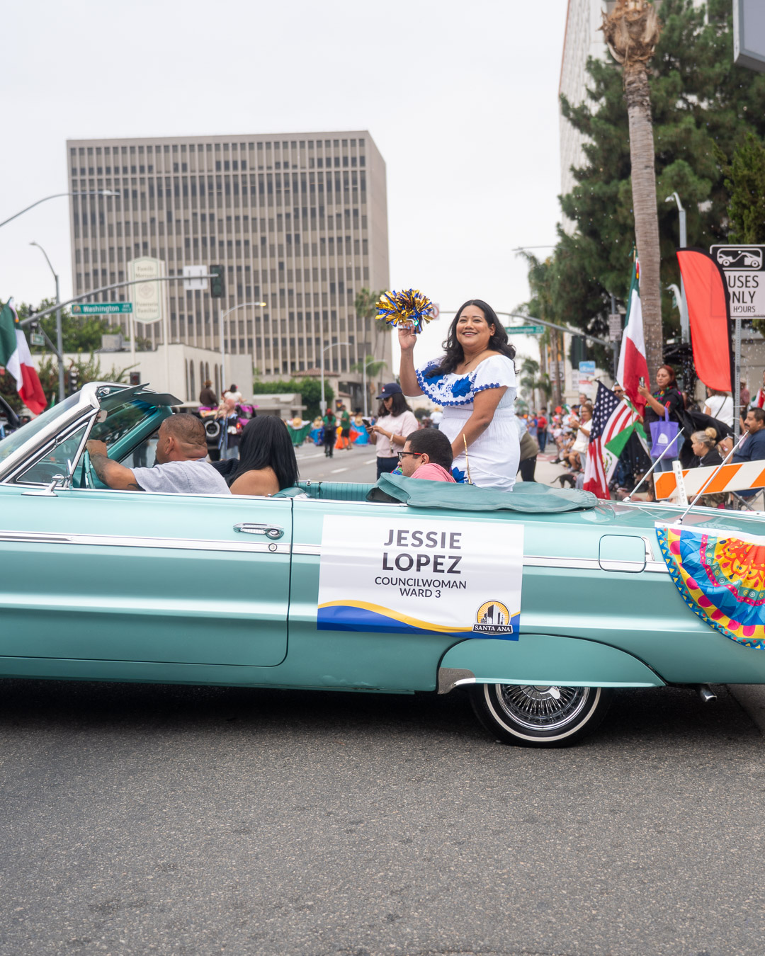 Woman standing in a vintage convertible car holding a bouquet, with a sign reading Jessie Lopez, in a parade with flags and spectators.