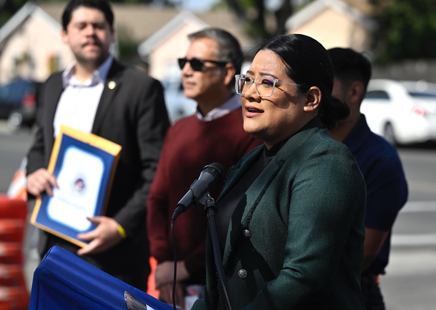 Woman speaking at a podium outdoors, wearing glasses and a dark green blazer, with three men behind her, one holding a framed document.