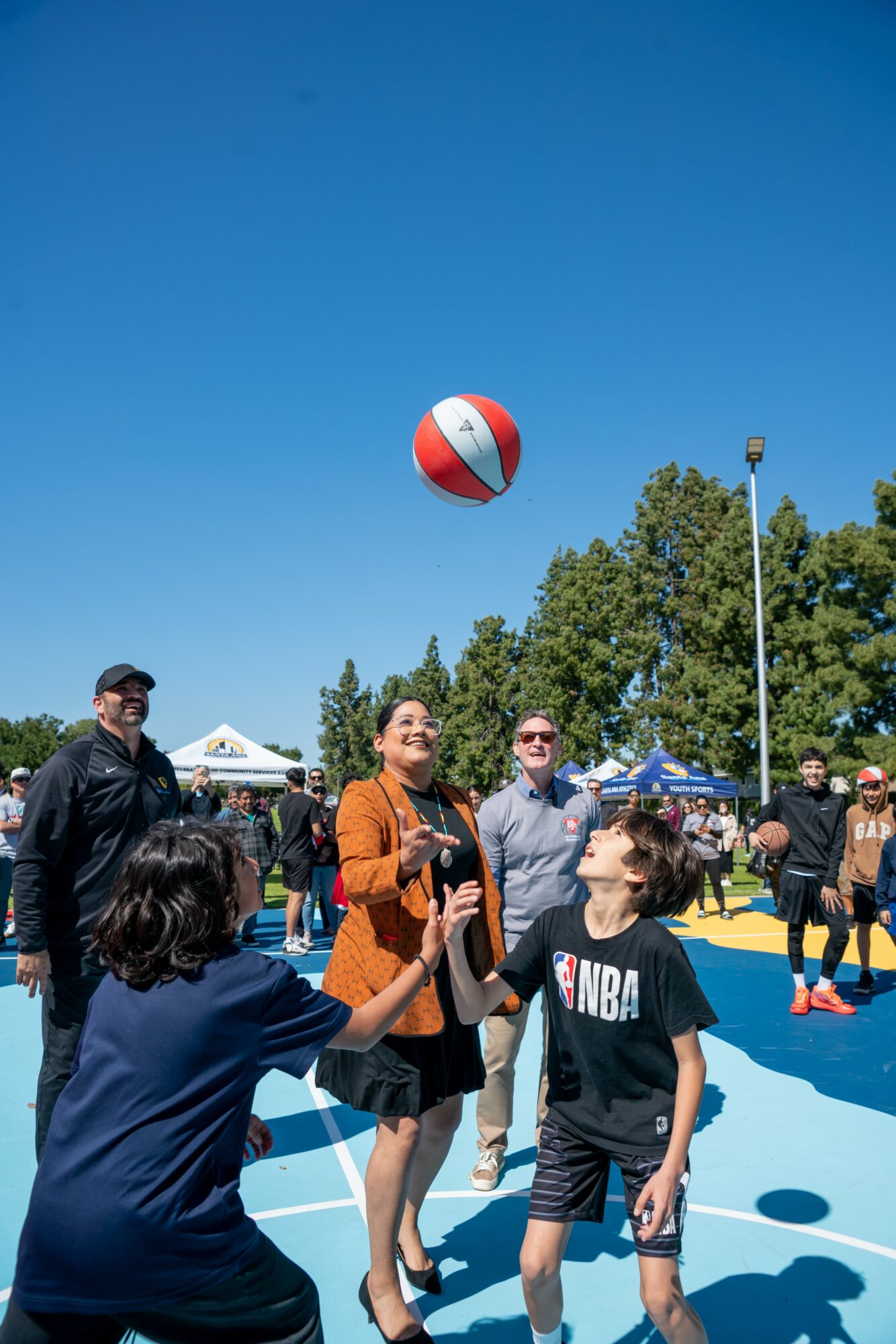 People playing basketball outdoors under a clear blue sky, with trees and a crowd in the background.