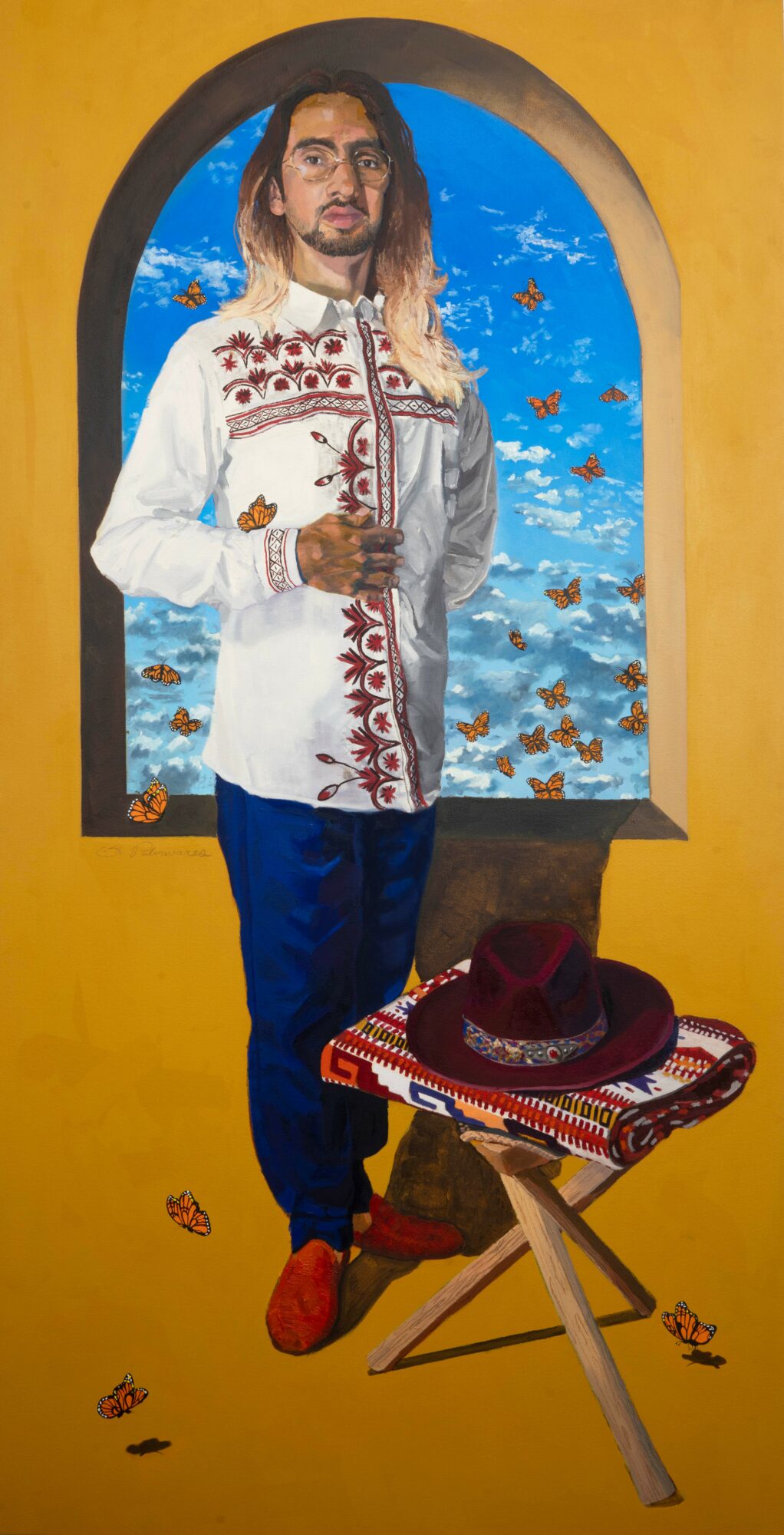 Person with long hair in a white embroidered shirt, standing indoors near a window with a blue sky and clouds, a hat on a table.