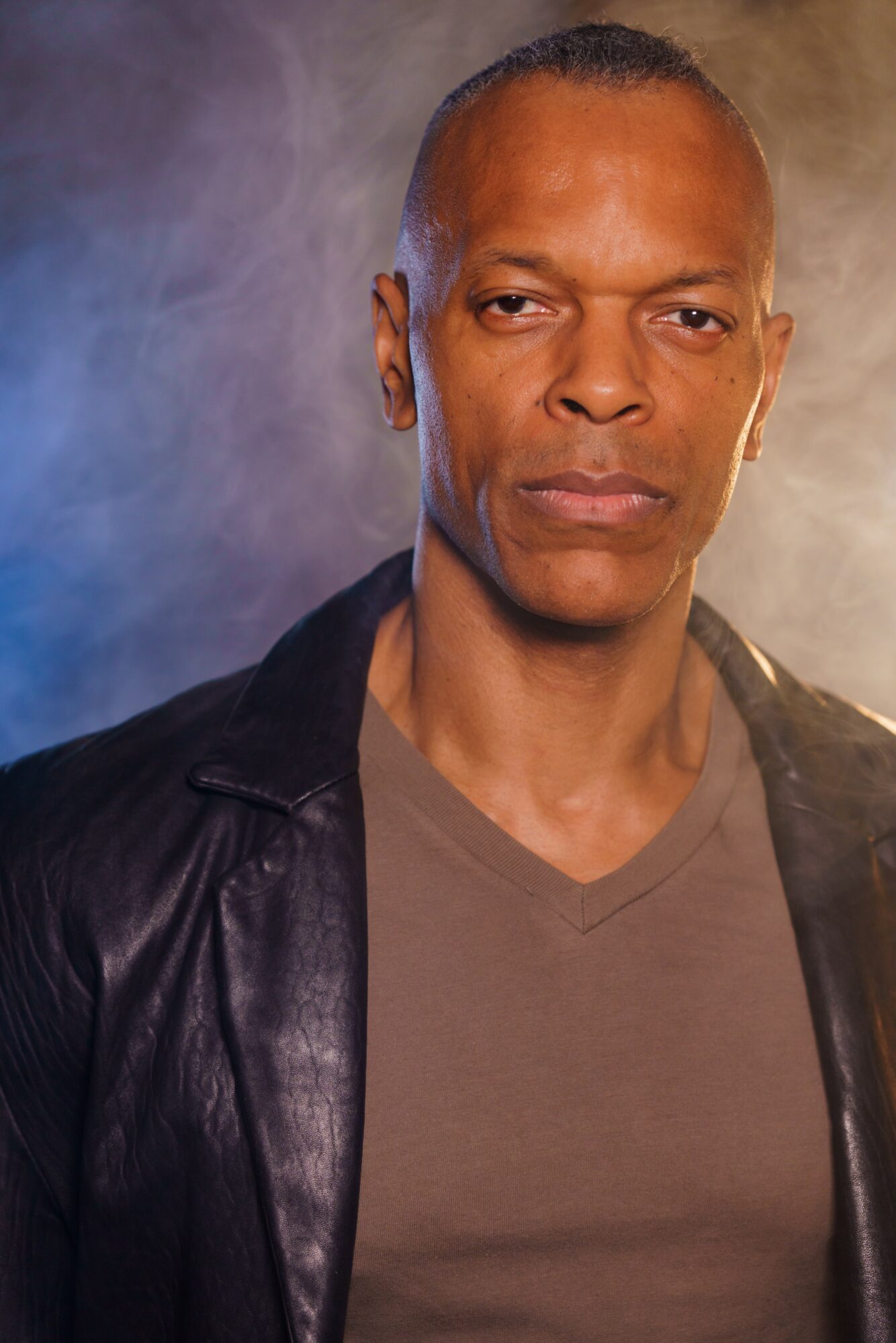 Man with short hair and serious expression wearing a leather jacket and gray shirt, against a smoky background.