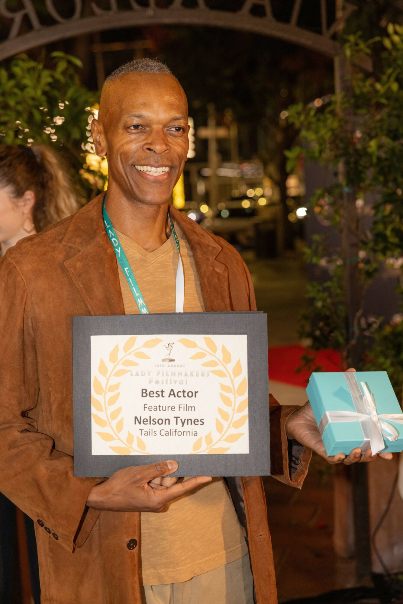 Man holding an award plaque and a gift box outdoors at night, smiling, with trees and lights in background.