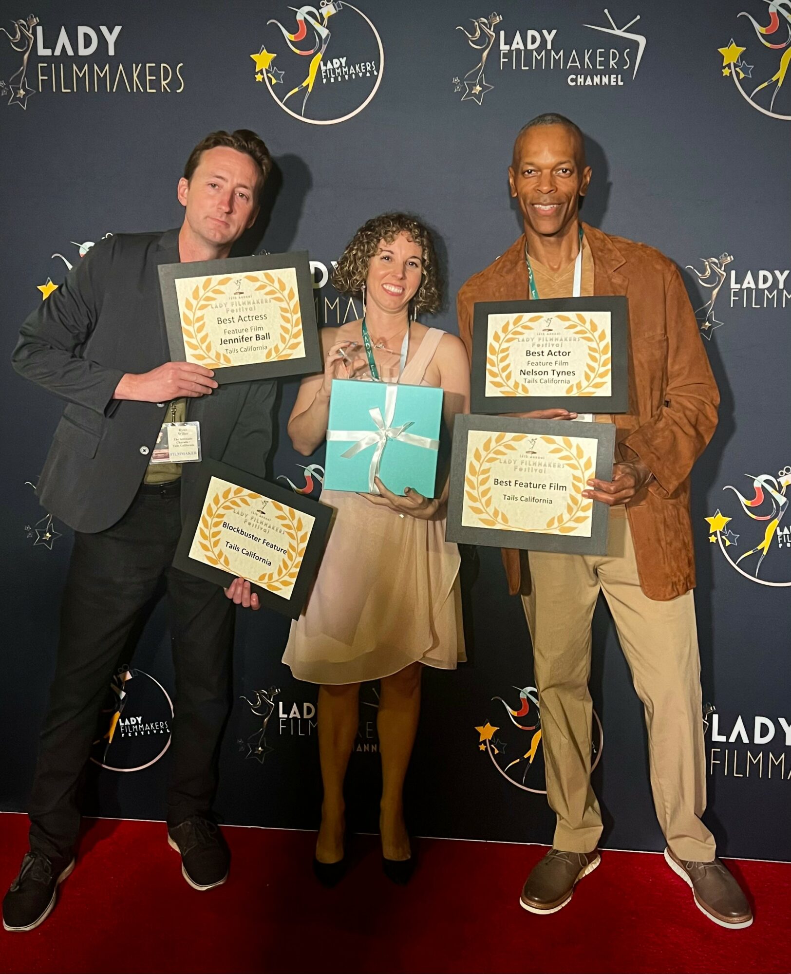Three people holding awards and a gift box, standing in front of a backdrop with logos, smiling at the camera.