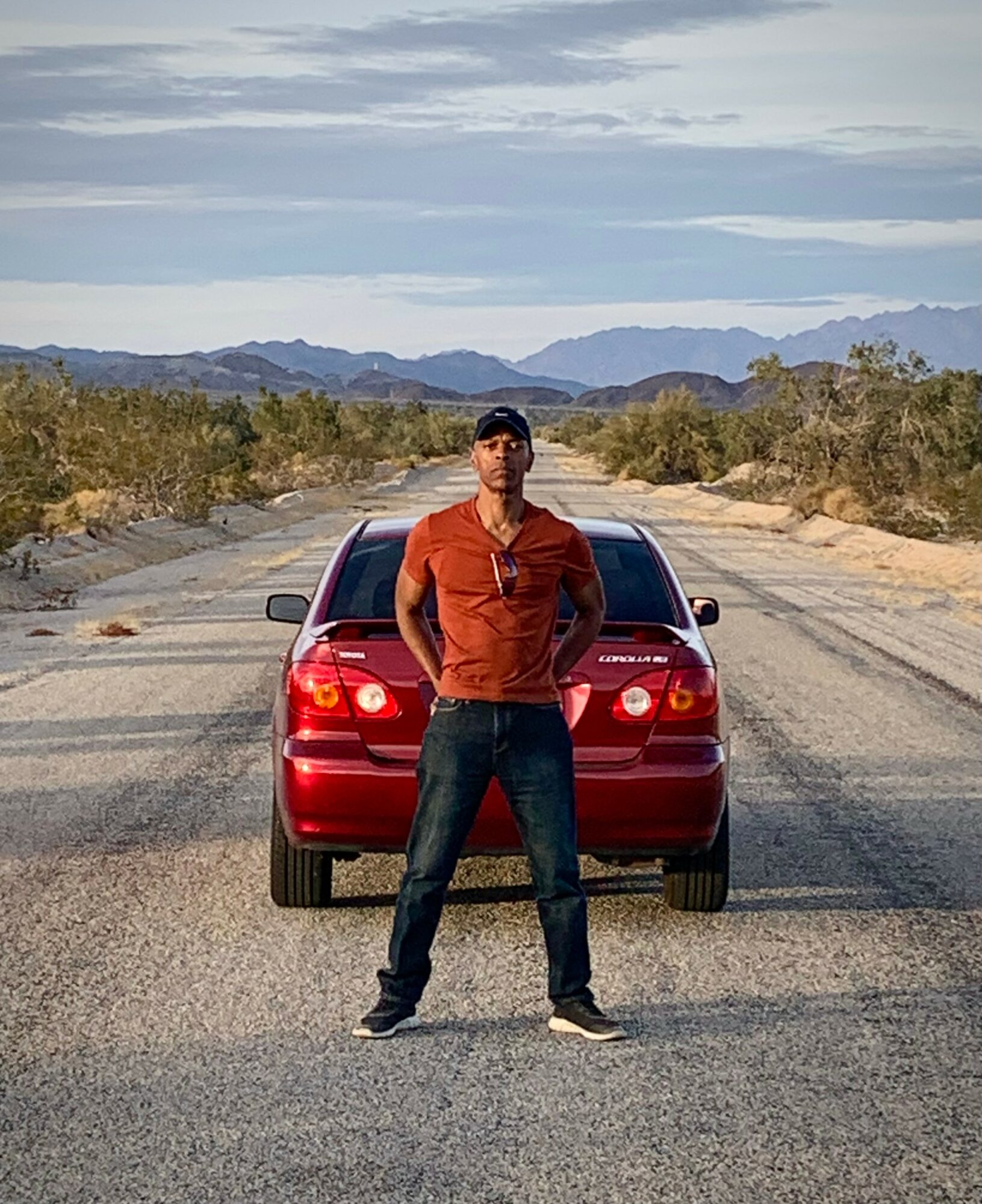 Person standing in front of a red car on a desert road with mountains in the background, under a cloudy sky.