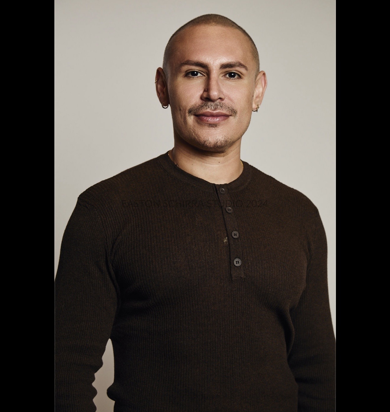 Man with a shaved head and light facial hair, wearing a dark long-sleeve shirt, smiling slightly, against a plain background.