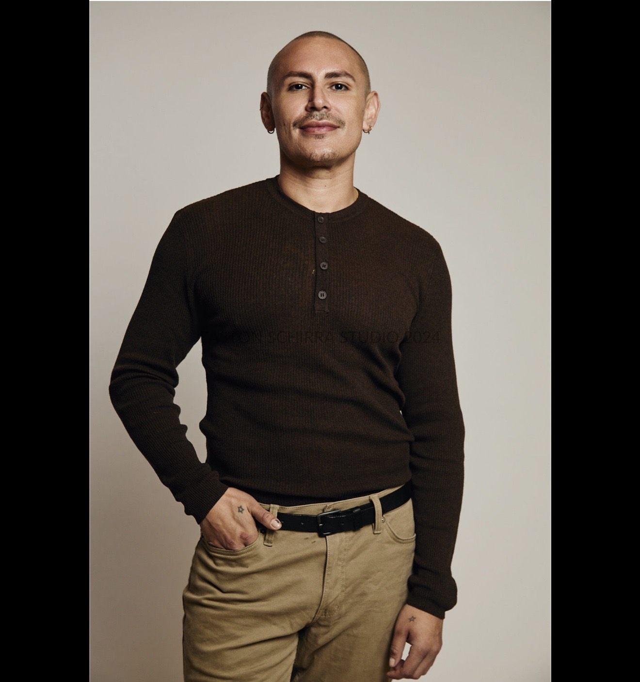 Person with a shaved head wearing a dark long-sleeve shirt and beige pants, standing against a plain background.