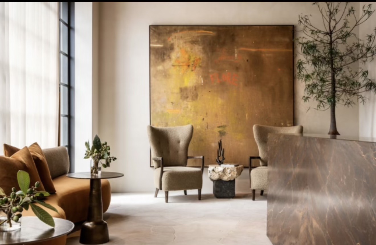 Living room with large abstract painting, two chairs, a small table, and plants, illuminated by natural light from windows.