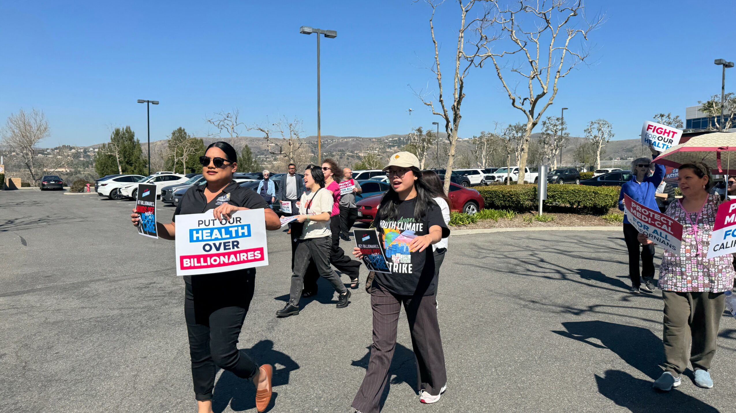 Group of people holding signs during a protest or rally outdoors, with parking lot and trees in background.
