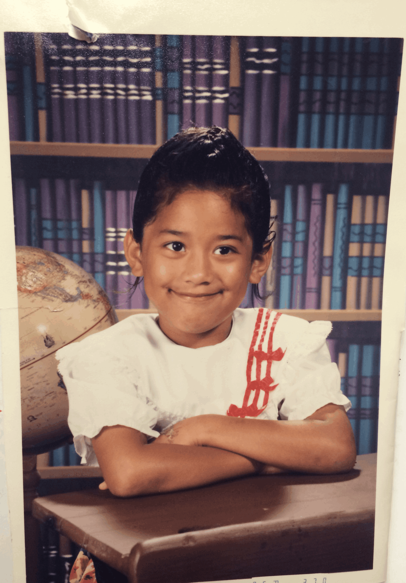 Child with short dark hair smiling, sitting at a desk with arms crossed, in front of a bookshelf filled with books.
