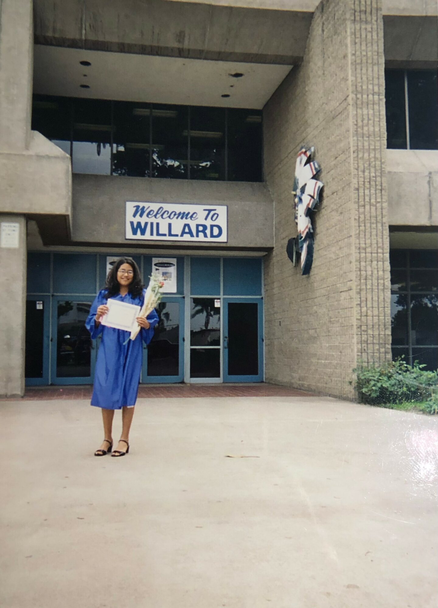 Person in blue graduation gown holding a diploma in front of a building with a sign that reads 'Welcome To WILLARD'.