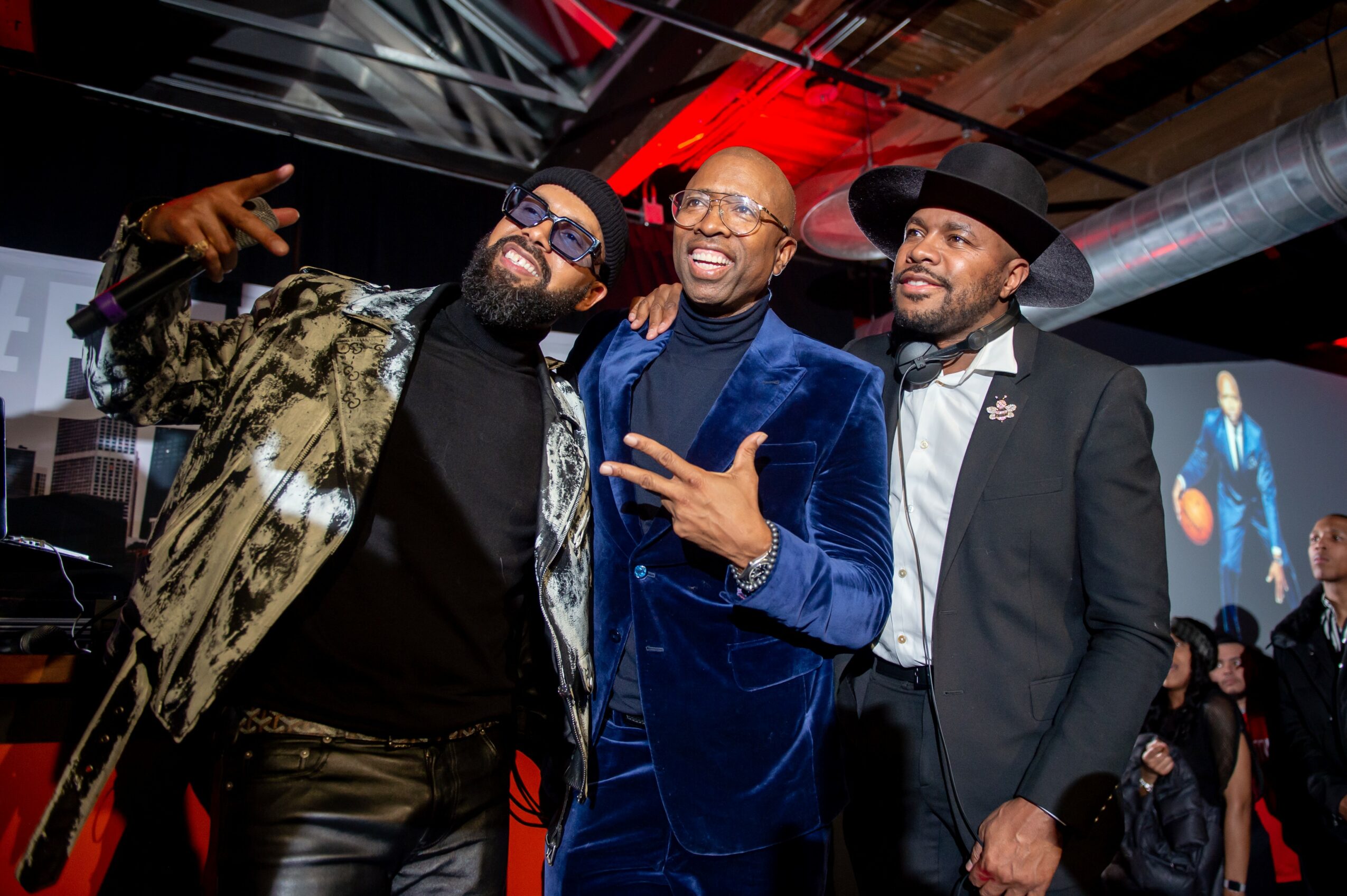 Three men smiling and posing together at an event, with a large screen and people in the background.
