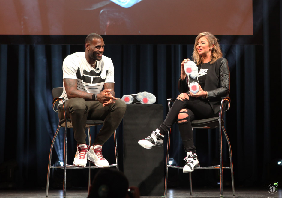 Two people sit on stage chairs, engaging in conversation with a black curtain background and a table between them.