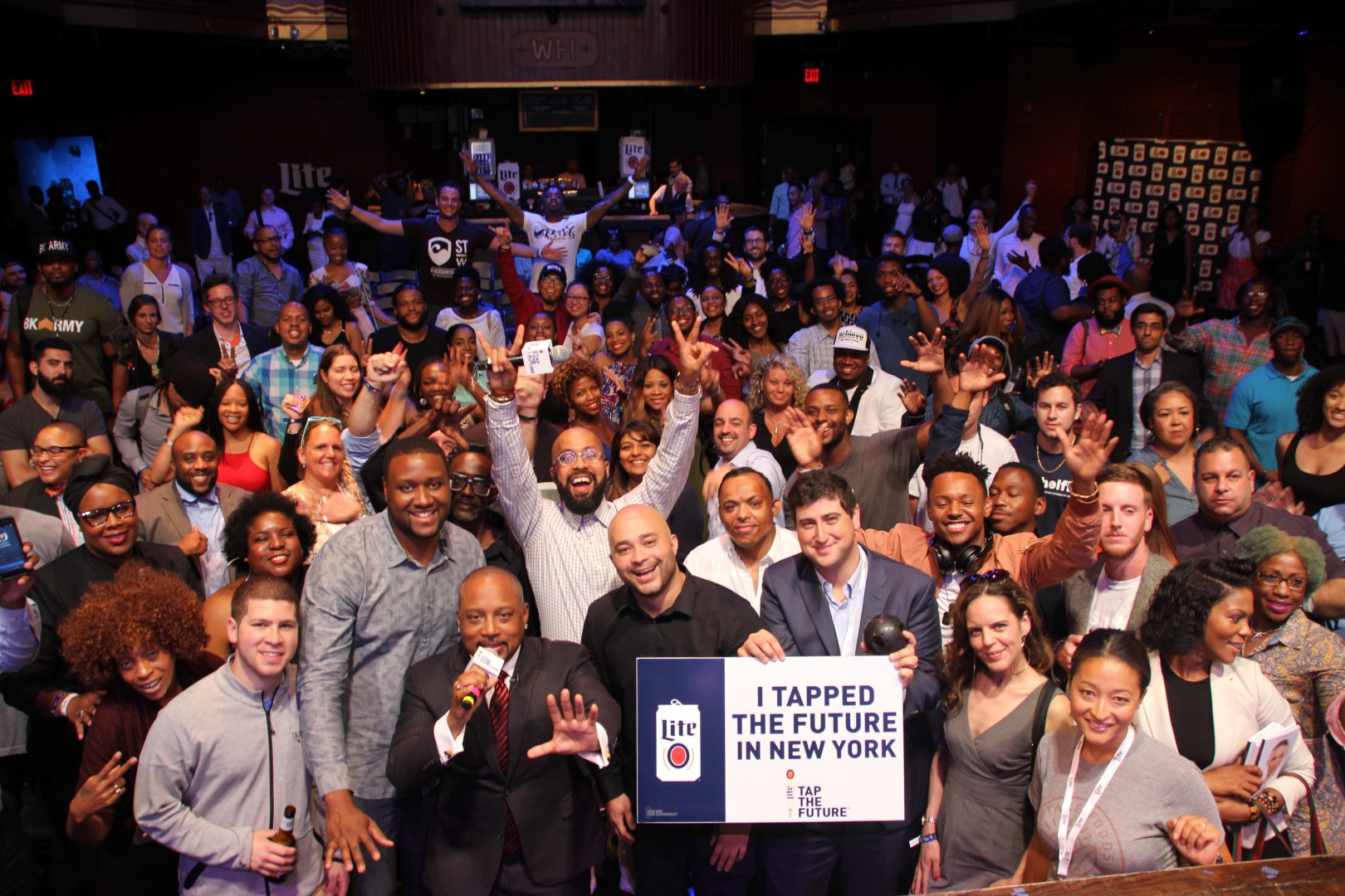 Large group of diverse people gathered indoors, smiling and waving, with a stage and screens in the background.