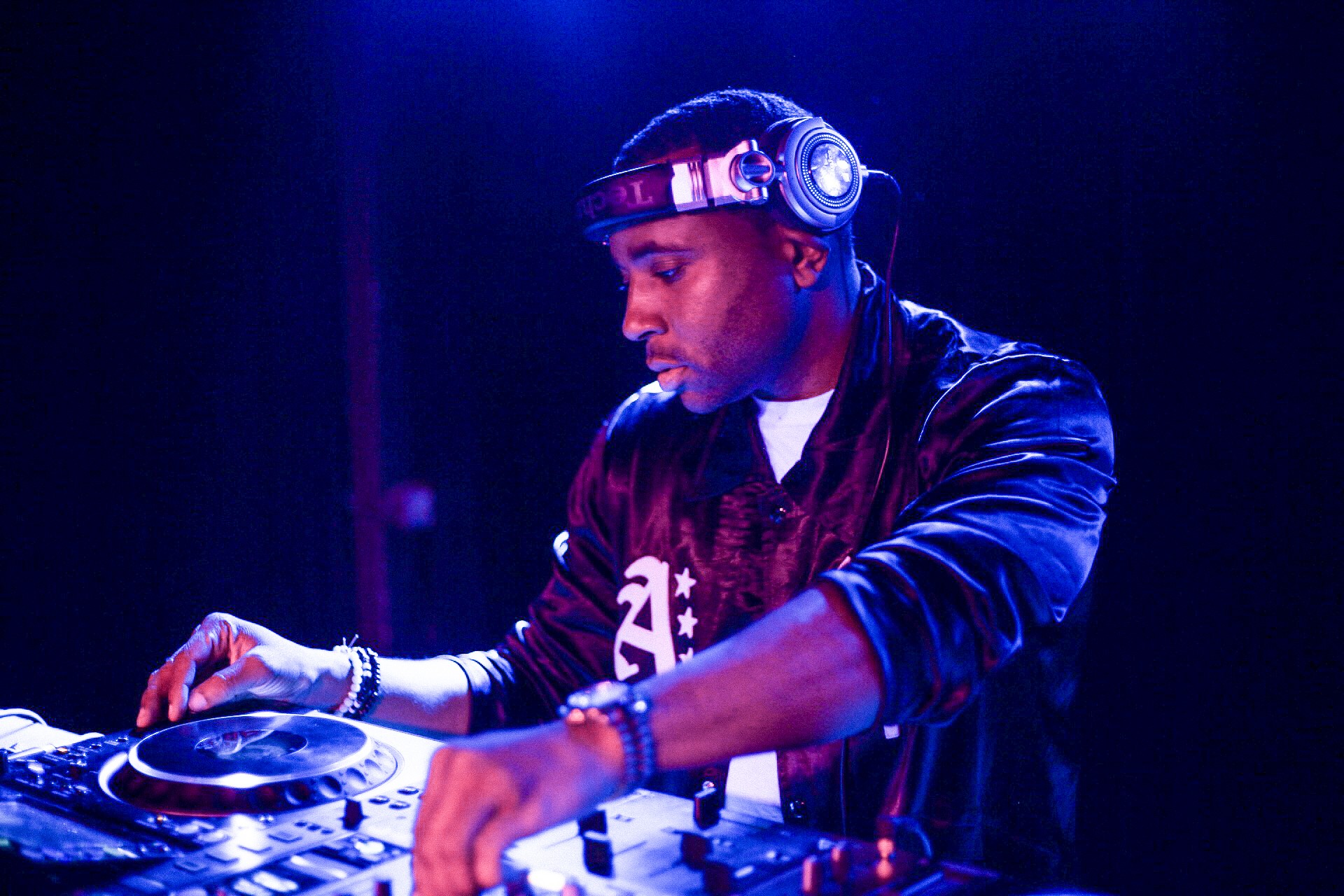 DJ wearing headphones and a jacket, focused on mixing music in a dark setting with blue lighting.