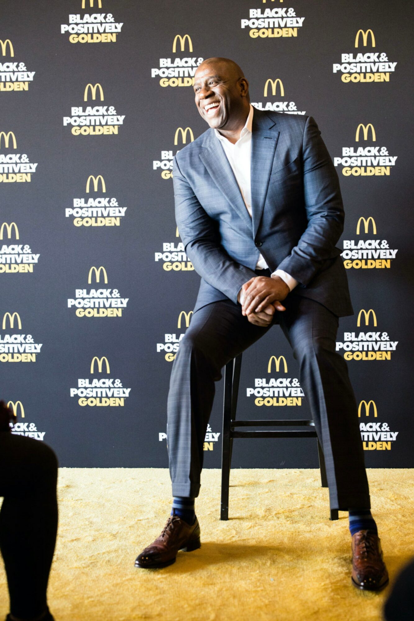 Man in a blue suit standing on a yellow carpet in front of a black backdrop with logos, smiling, hands clasped.