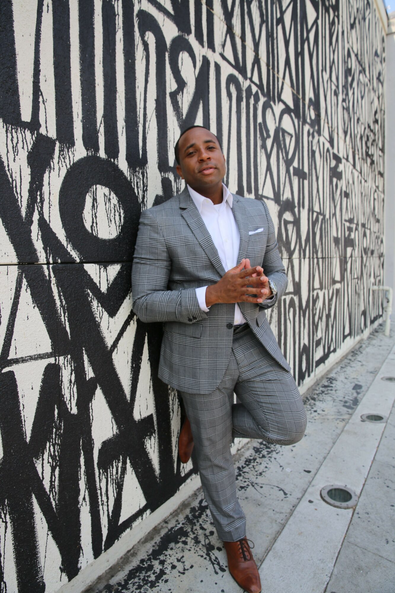 Man in gray suit holding a phone, standing against a graffiti-covered wall.
