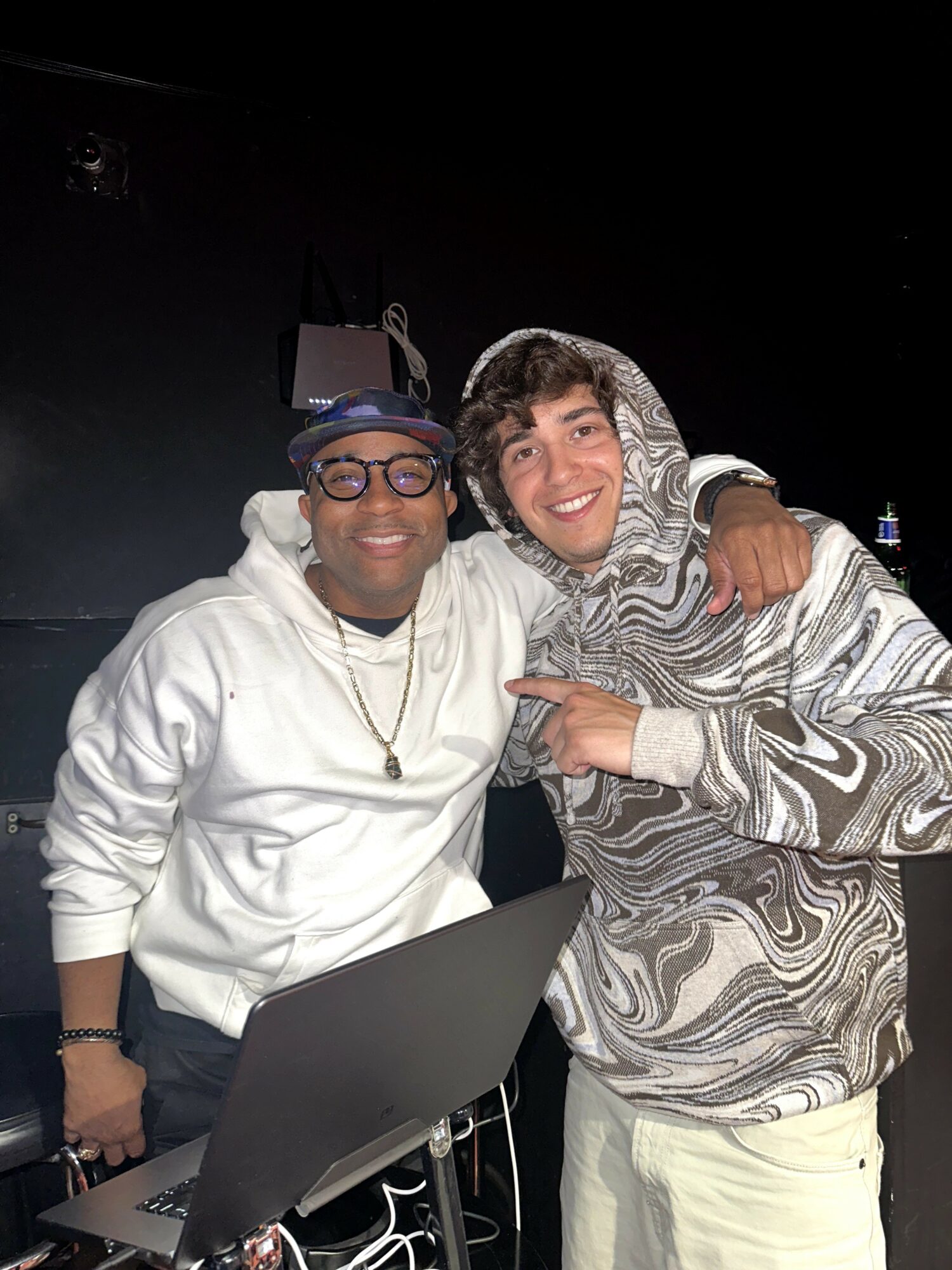 Two smiling people, one wearing glasses and a hoodie, the other in a patterned hoodie, hugging in a dark setting.