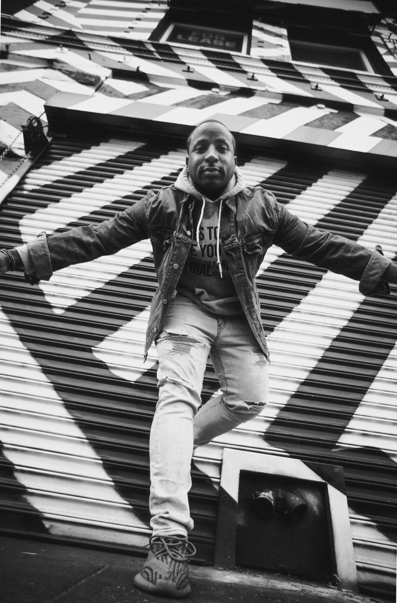 Person standing with arms outstretched in front of a geometric patterned wall, smiling, in black and white.