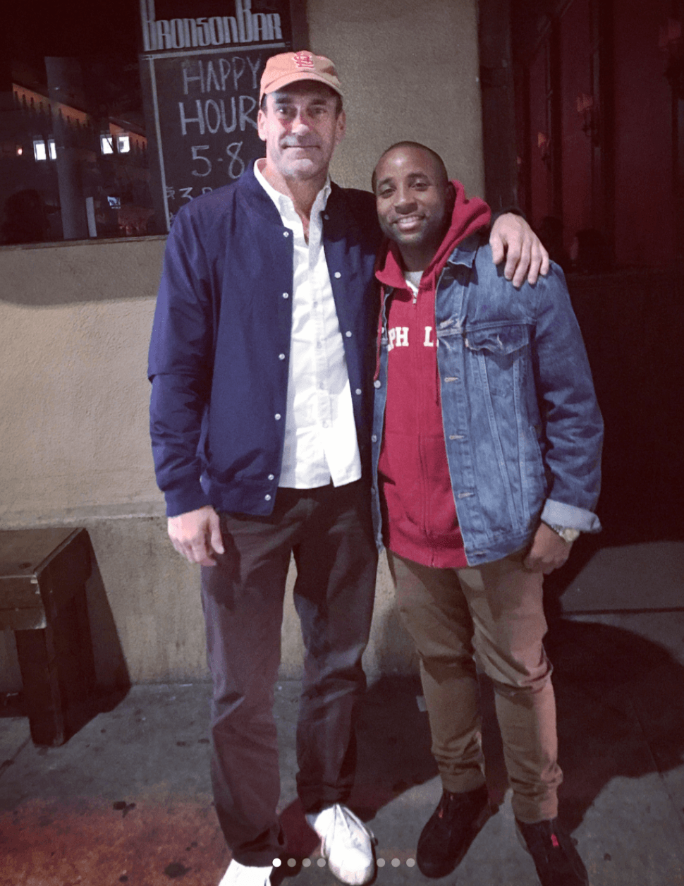 Two men standing outside at night, one with arm around the other, smiling, with a sign and window behind them.