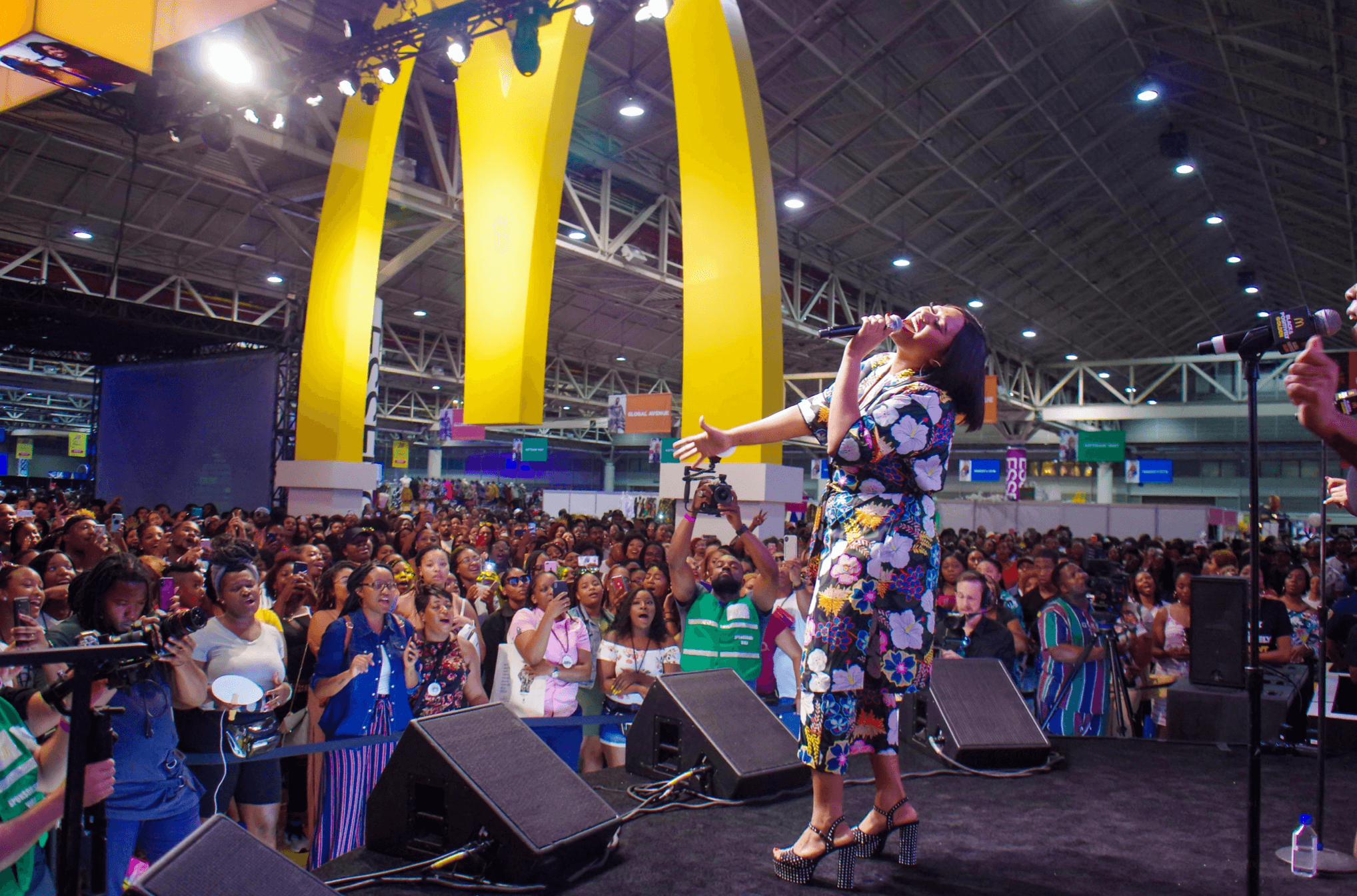 Performer on stage with a floral dress and headscarf, audience in a large indoor event space, bright yellow arches overhead.