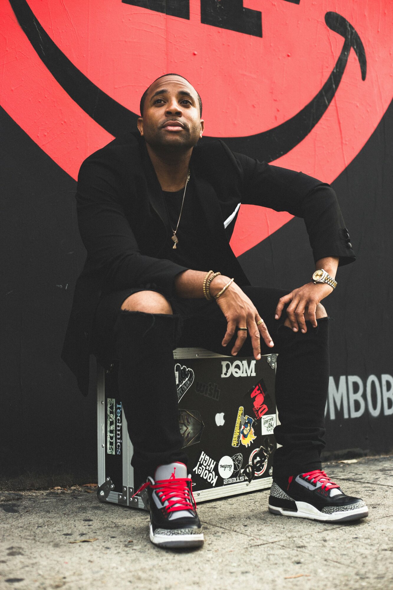Man sitting on a decorated box in front of a large red smiley face mural, wearing black clothing and sneakers.