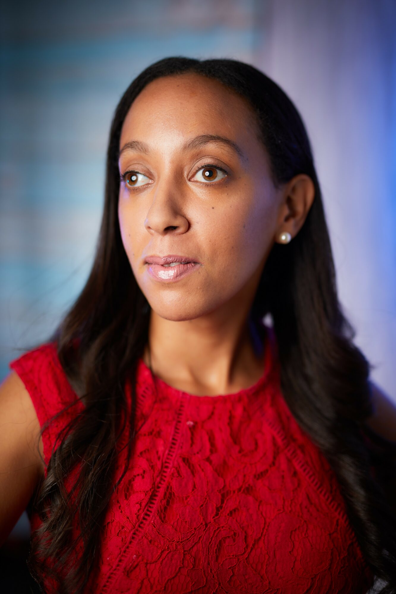 Woman with long dark hair wearing a red sleeveless top, looking to the side, with a blurred background.