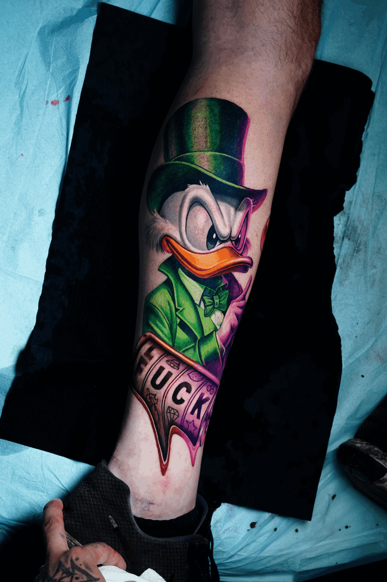 Colorful tattoo of a cartoon duck with a green suit and purple tie on person's leg.